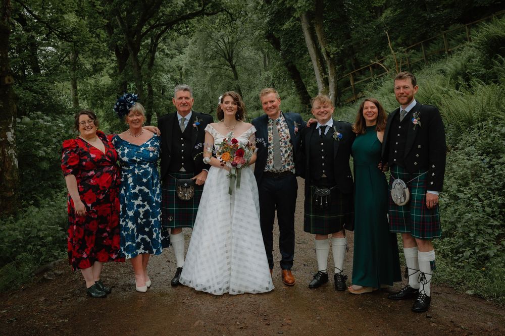 Wedding party posing on forest path, bride in lace gown with groom and guests in Scottish kilts and formal attire.