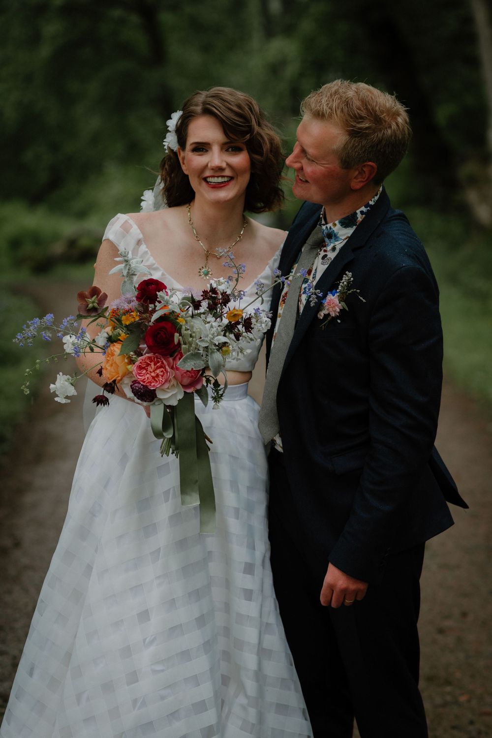 Bride holding colorful bouquet stands with groom in navy suit on a forest path, both smiling on their wedding day.
