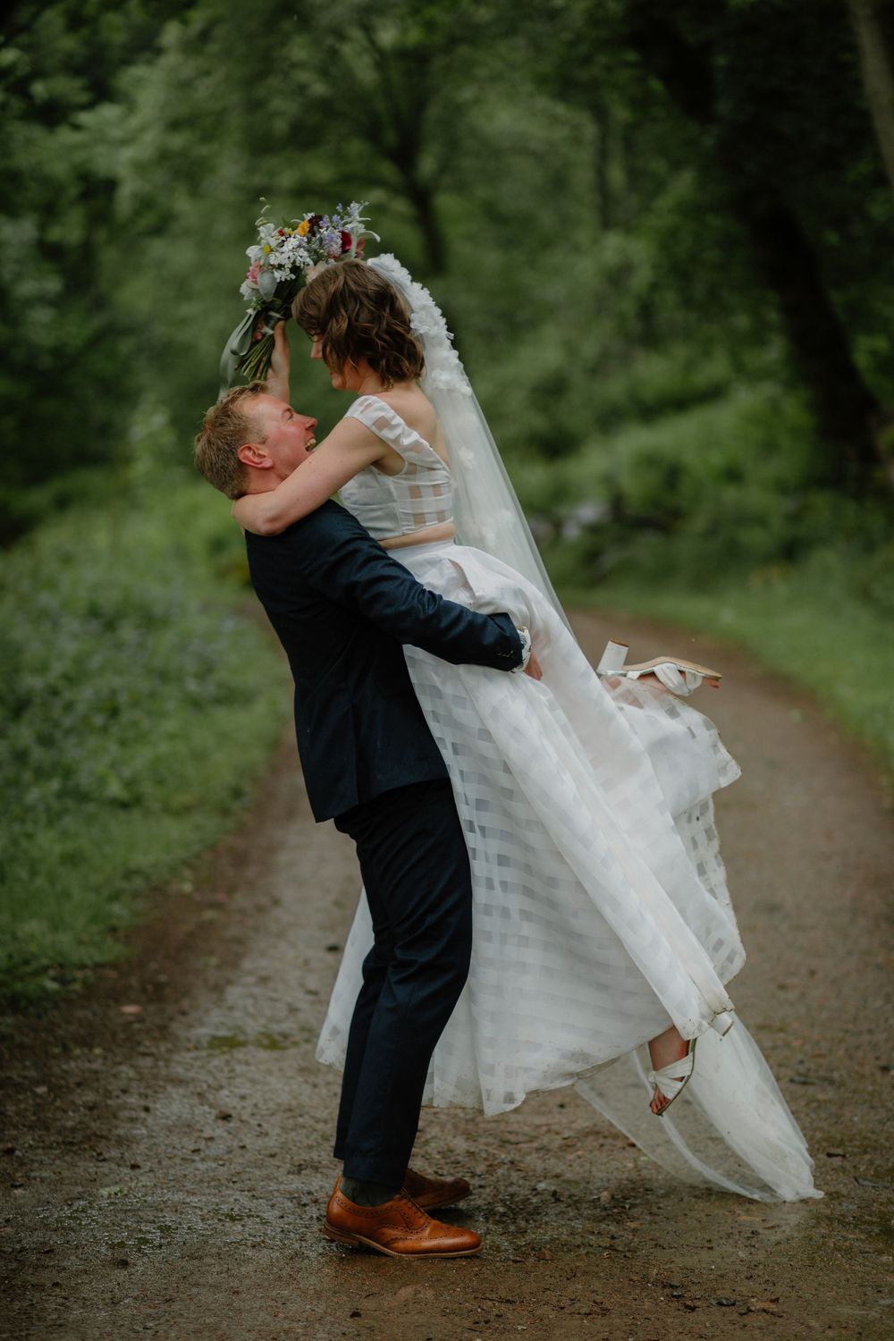 Groom lifting bride in white gown and veil on a lush green forest path during outdoor wedding celebration.