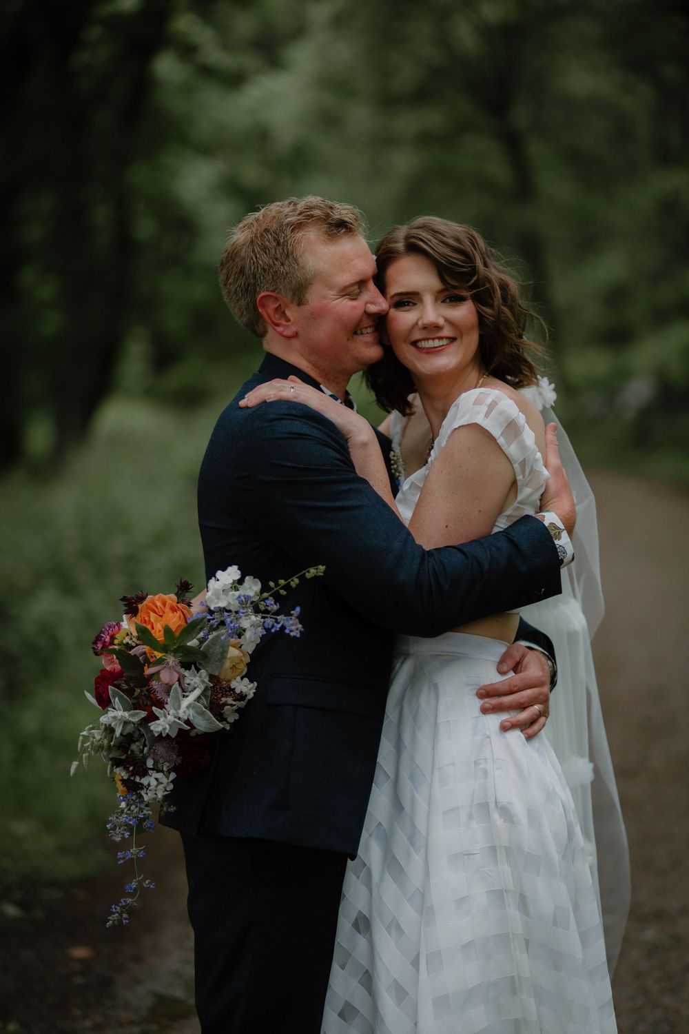 Bride and groom embrace on a forest path, she holds a colorful wildflower bouquet in a white dress.