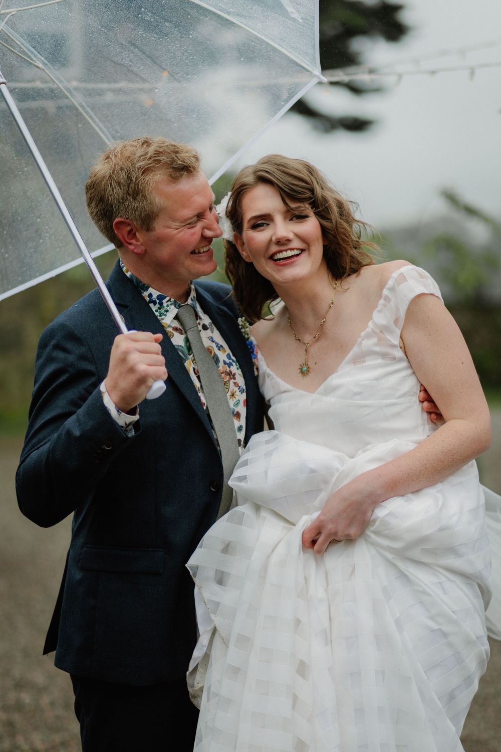 A joyful bride and groom share a laugh under a clear umbrella on their rainy wedding day.