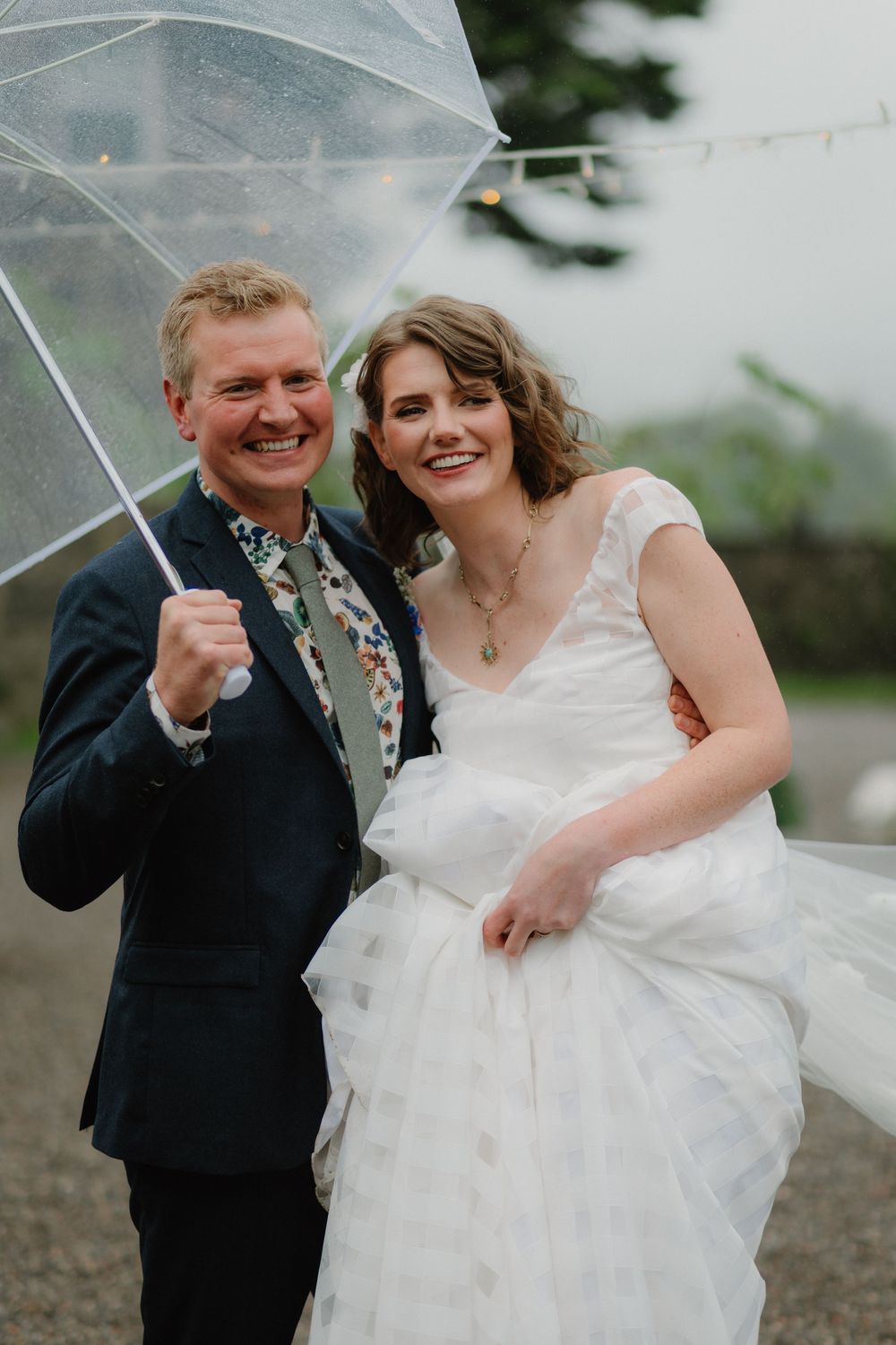 Smiling bride and groom share a clear umbrella on their rainy wedding day, bride holding white gown.