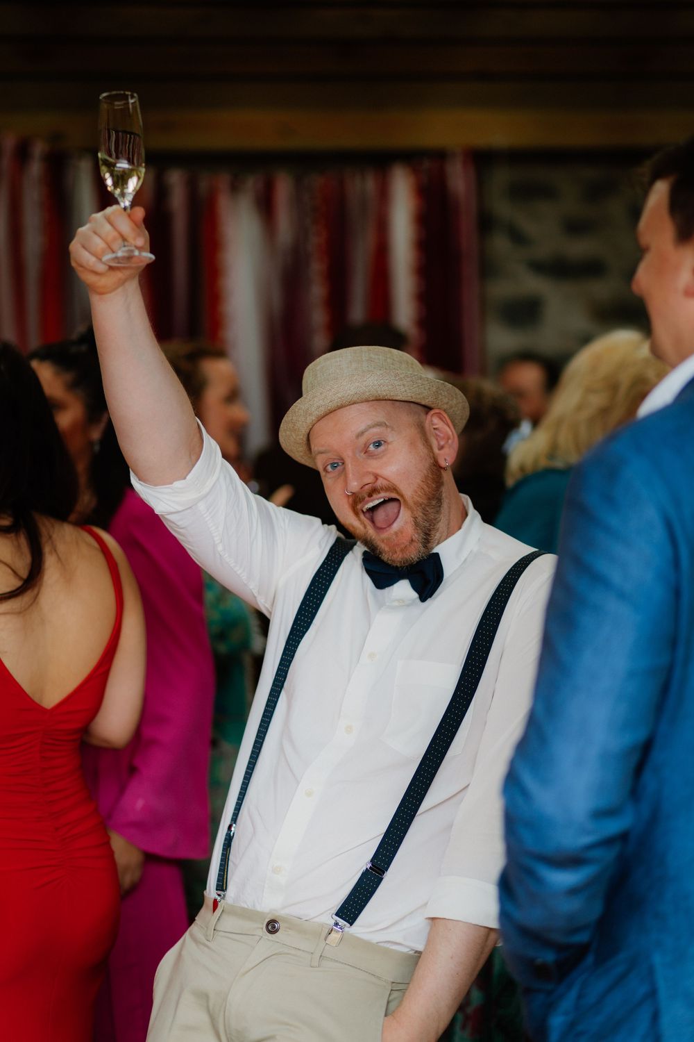 Man in suspenders and bow tie raises champagne glass at a festive celebration, wearing a tan hat and joyful expression.