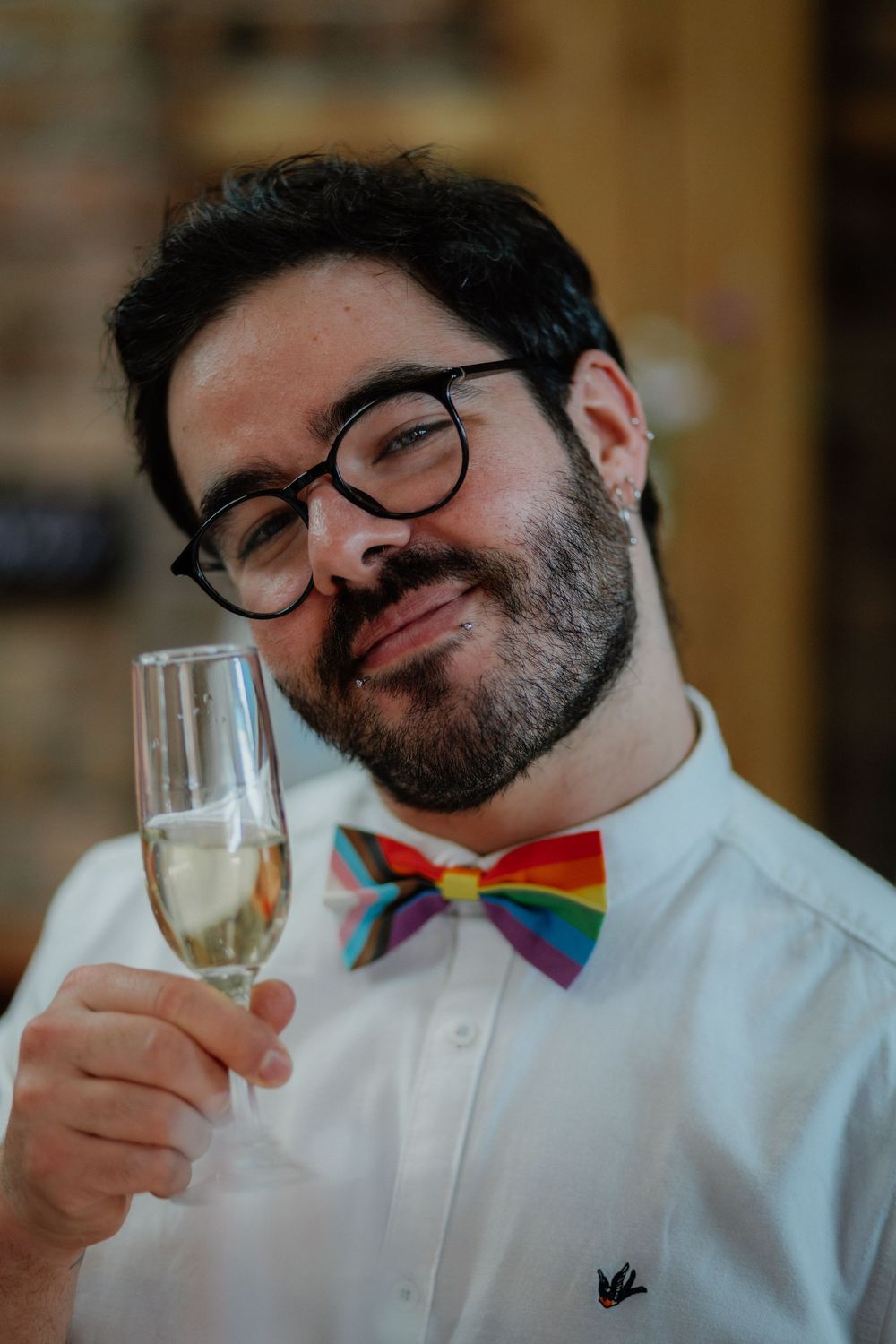 Man with glasses and rainbow bow tie holding champagne flute, smiling confidently at the camera.