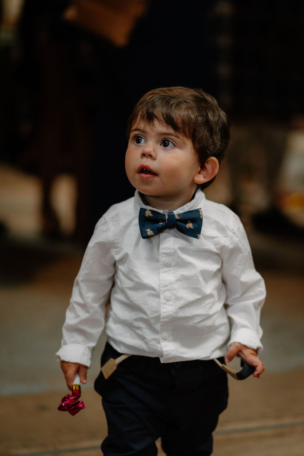 Adorable toddler boy in white shirt, navy bow tie, and suspenders looking up curiously at a formal event.