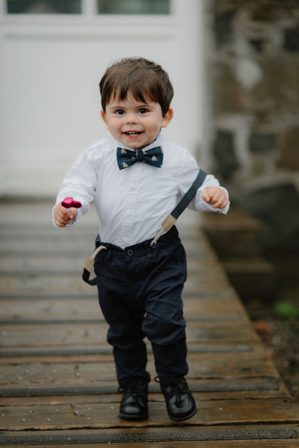 Adorable toddler boy in white shirt, bow tie, suspenders, and black pants smiling on a wooden deck.