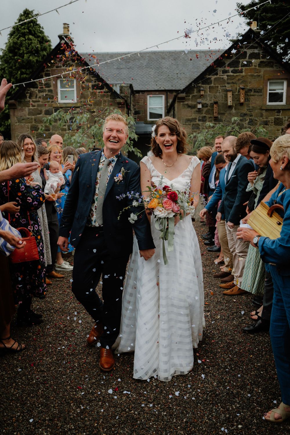 Happy newlyweds walk through confetti shower at outdoor wedding ceremony, bride holding colorful bouquet.