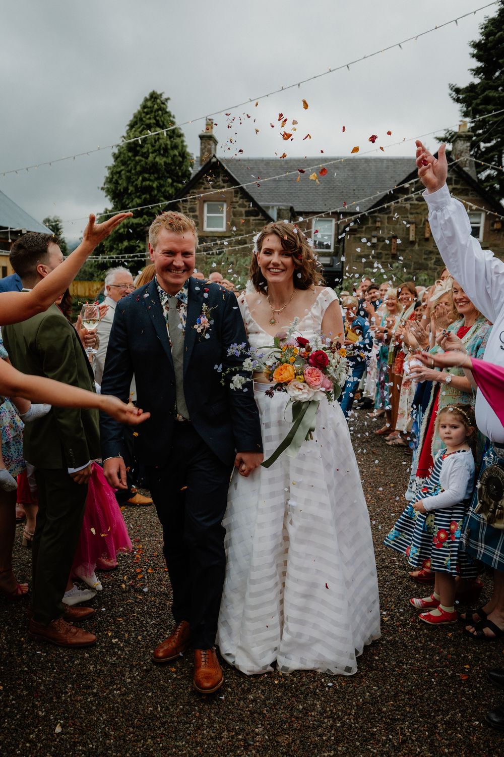 Bride and groom walk through colorful confetti shower surrounded by cheering wedding guests outside a stone building.