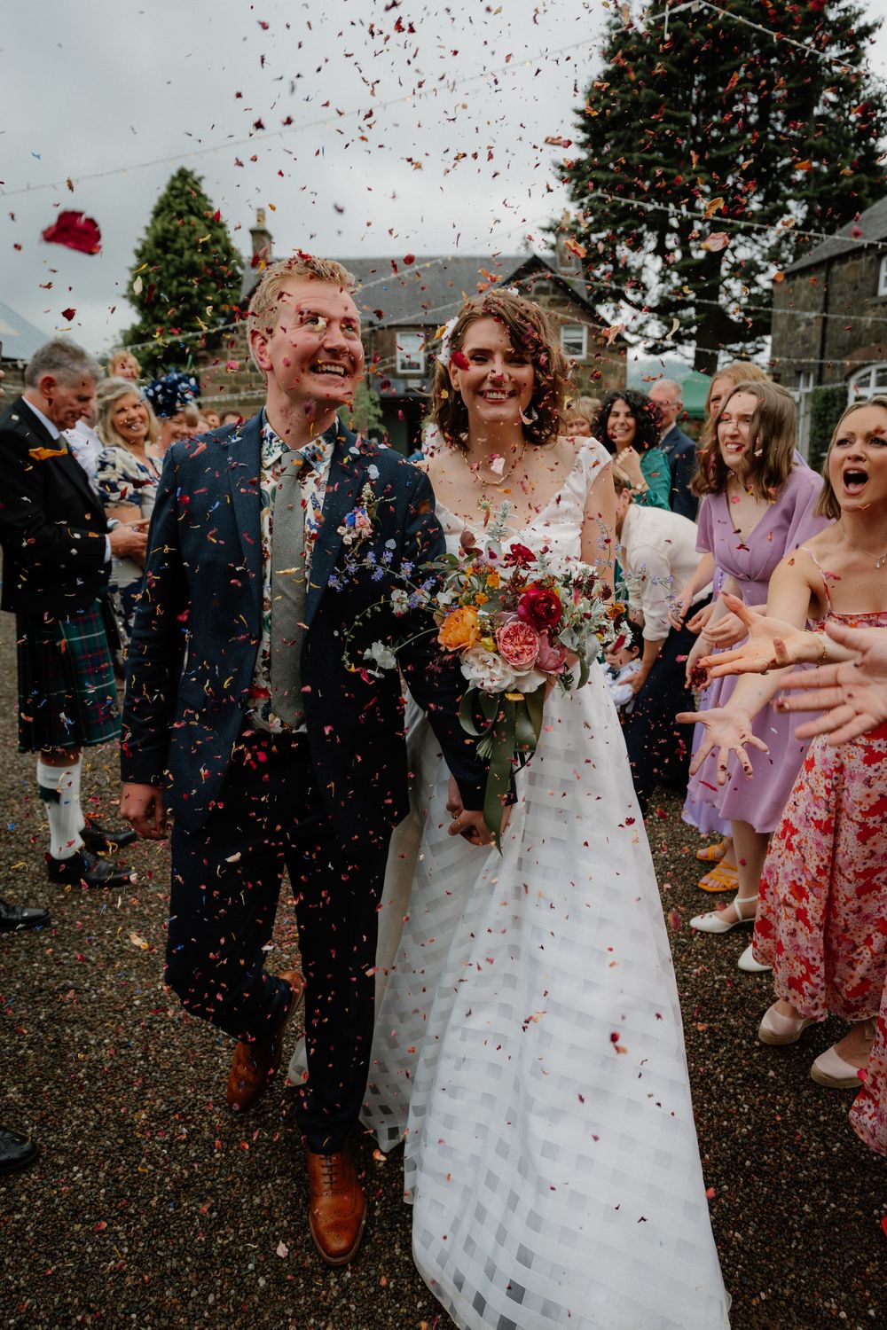 Happy newlyweds smile as vibrant confetti rains down during their outdoor wedding celebration with guests cheering.