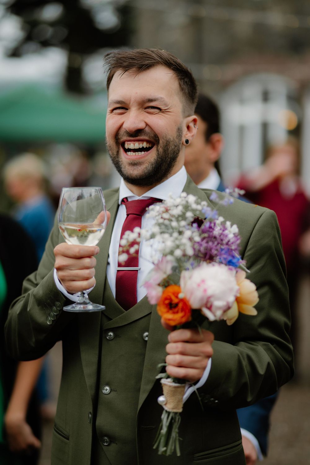 Laughing groom in green suit holding wine glass and colorful bridal bouquet at outdoor wedding celebration.