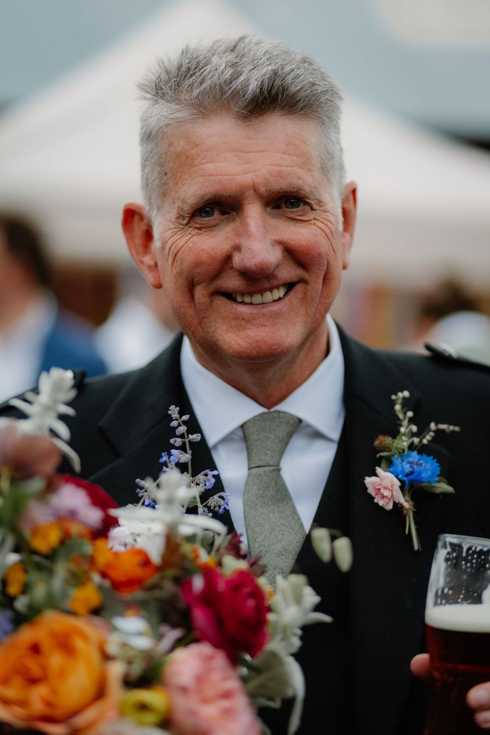 Smiling older gentleman in dark suit with floral boutonniere holds colorful bouquet and beer at a wedding reception.