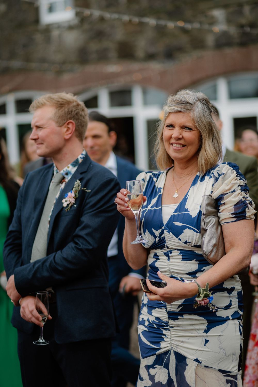 A smiling woman in a blue floral dress holds a champagne glass at an outdoor wedding reception.