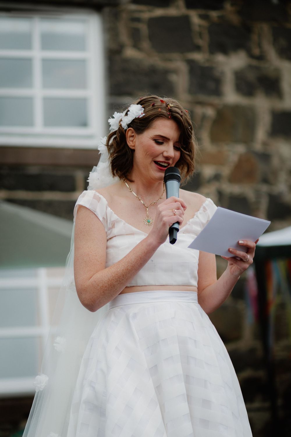 Bride in white dress reads vows into microphone at outdoor wedding ceremony.