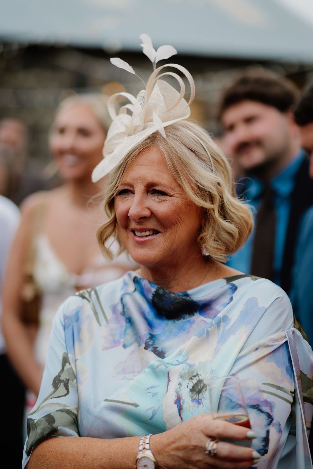 Elegant woman in floral dress and fascinator hat smiling at a wedding reception outdoor event.