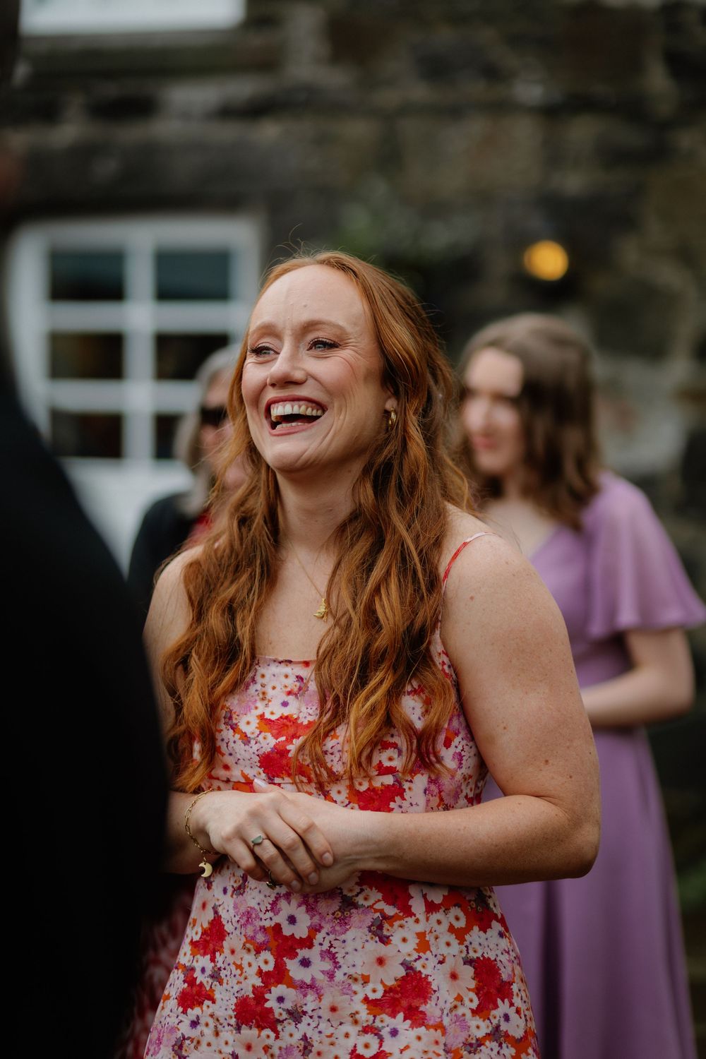 Laughing woman with long red hair wearing a floral dress at an outdoor wedding celebration.