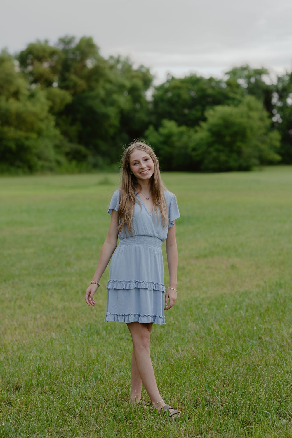 A smiling young girl in a light blue ruffled dress stands barefoot in a green grassy field with trees in the background.