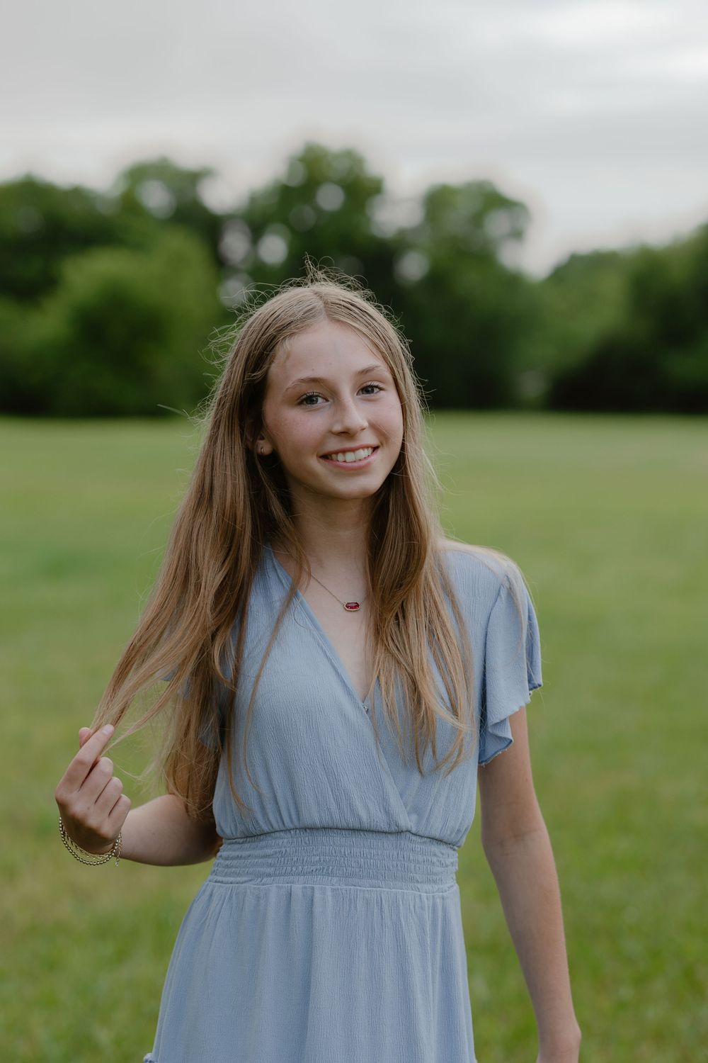 A smiling teenage girl in a light blue dress stands in a green field, her long blonde hair flowing naturally.