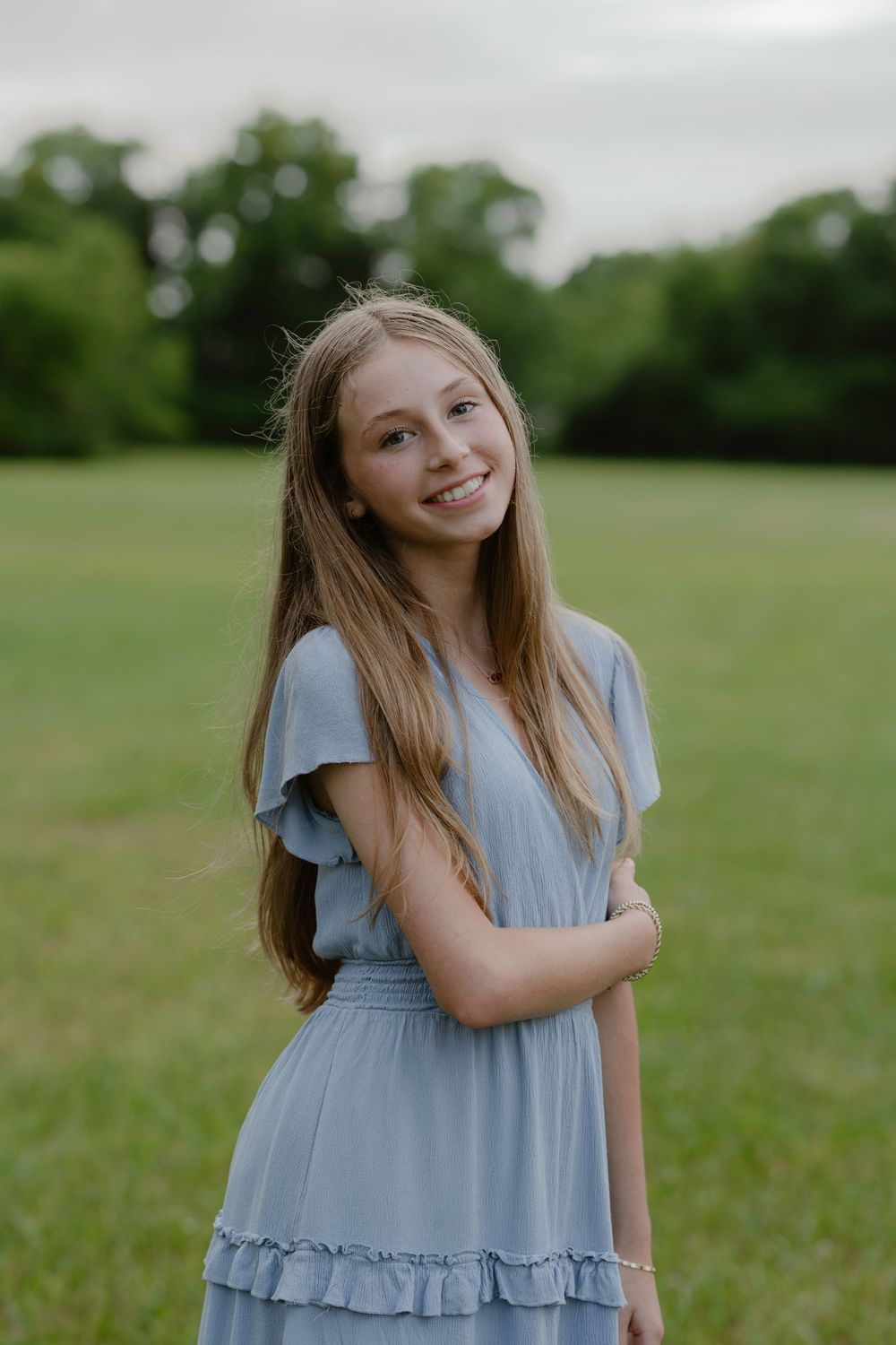 A smiling teenage girl in a light blue ruffled dress stands in a green field with trees in the background.