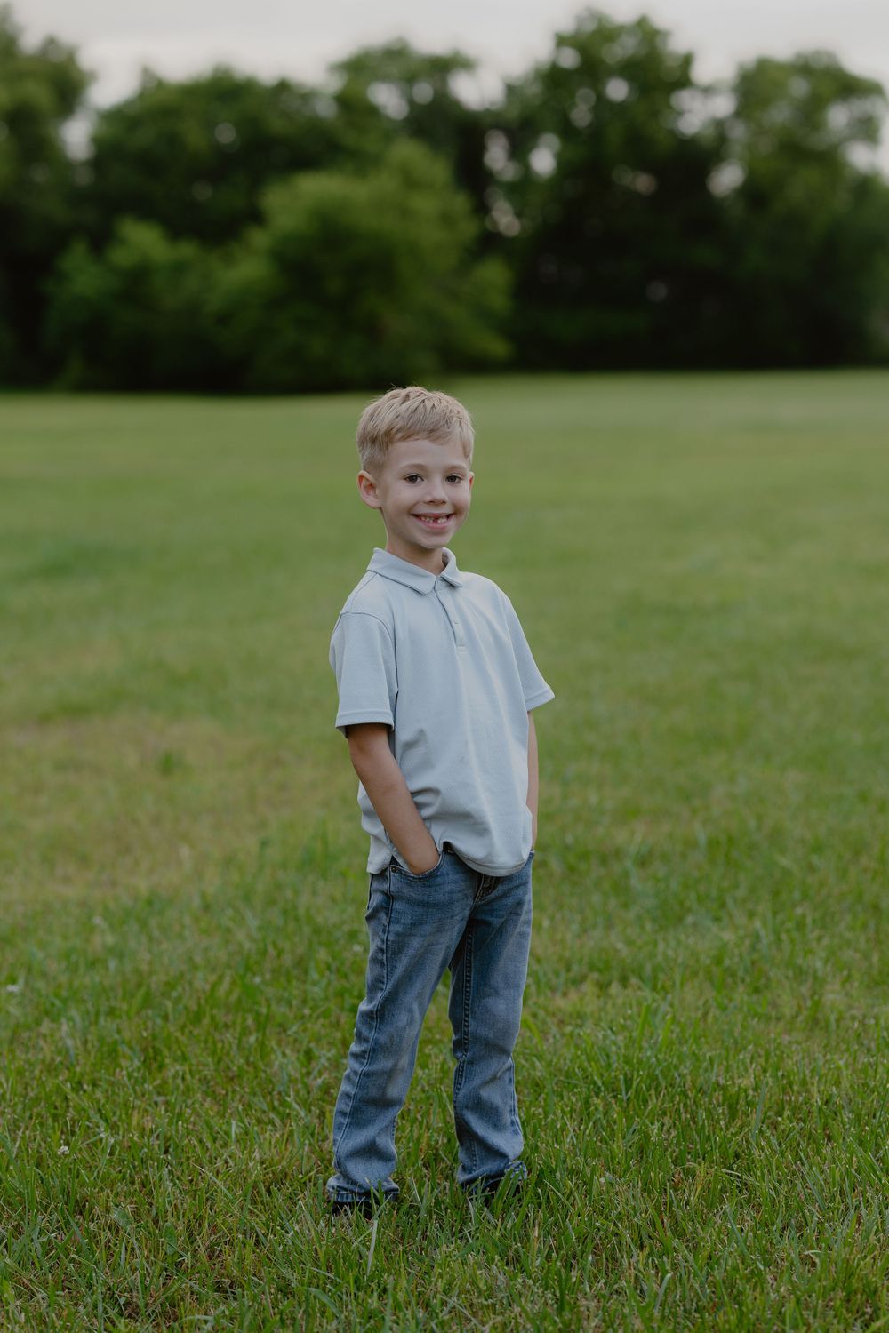 Young boy in light blue polo shirt and jeans smiling while standing in a green grassy field with trees in background.