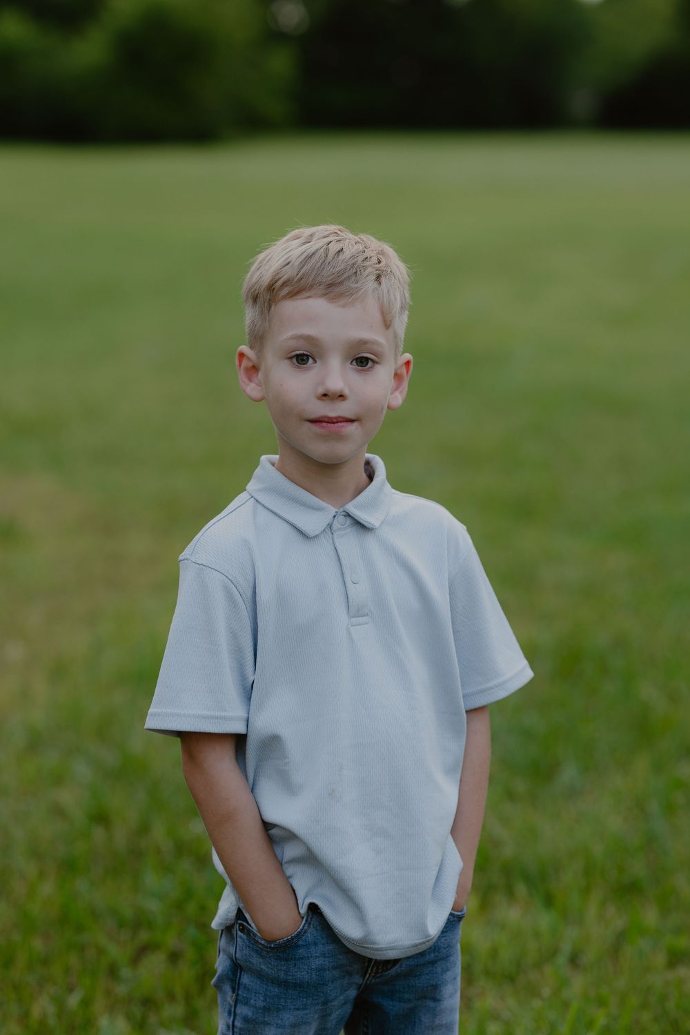 A young blonde boy in a light blue polo shirt stands outdoors on a green grassy field, smiling at the camera.