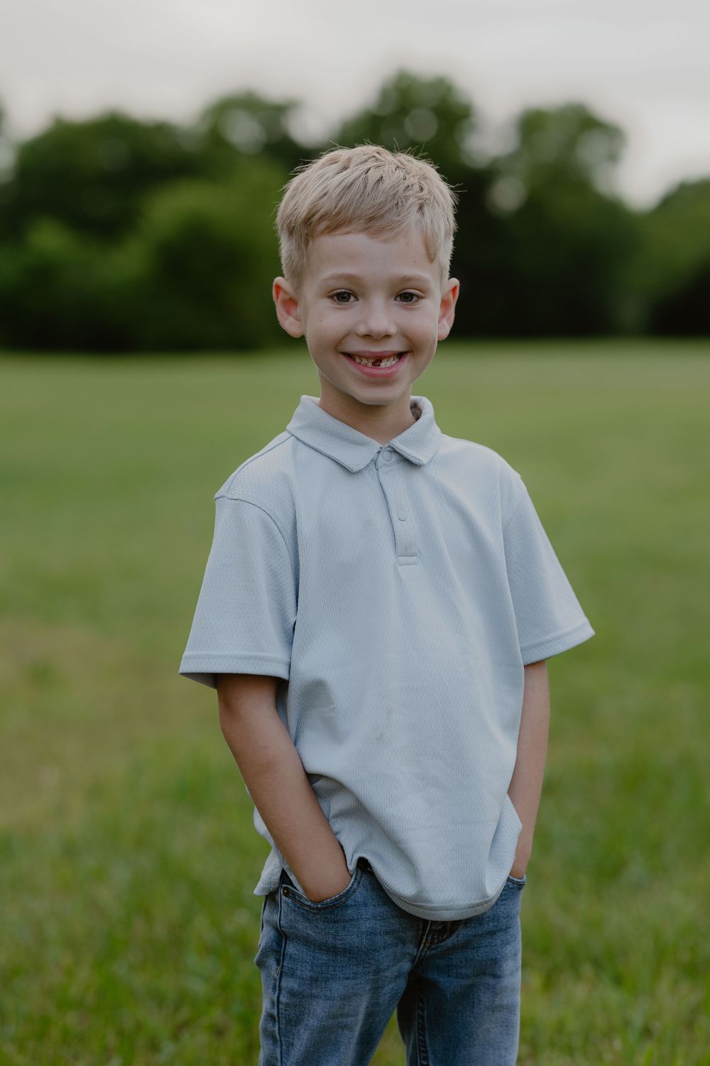 Smiling blonde boy in light blue polo shirt and jeans standing in a green field with trees in background.