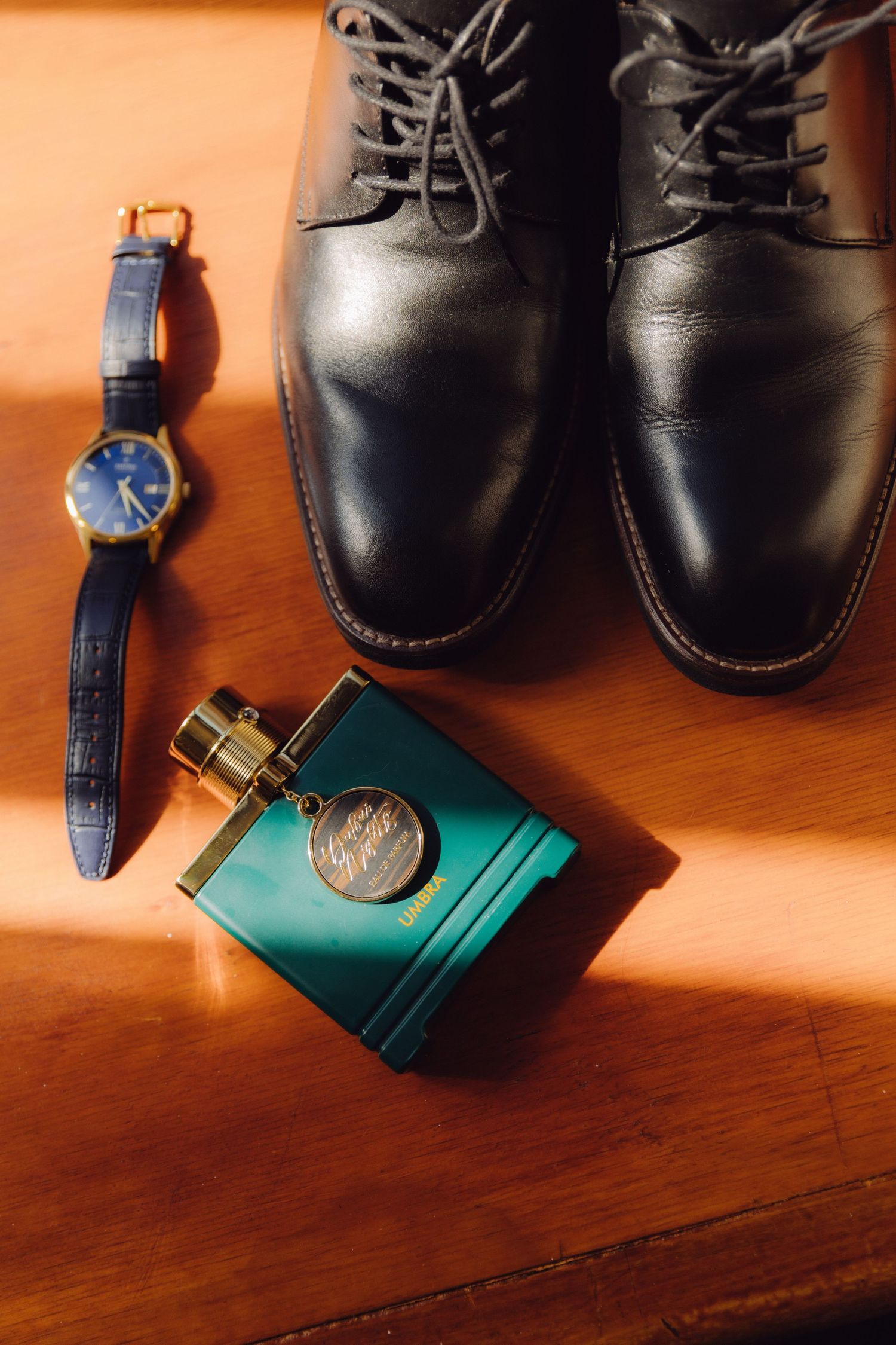 Groom accessories flat lay featuring black leather dress shoes, navy watch, and teal cologne bottle.