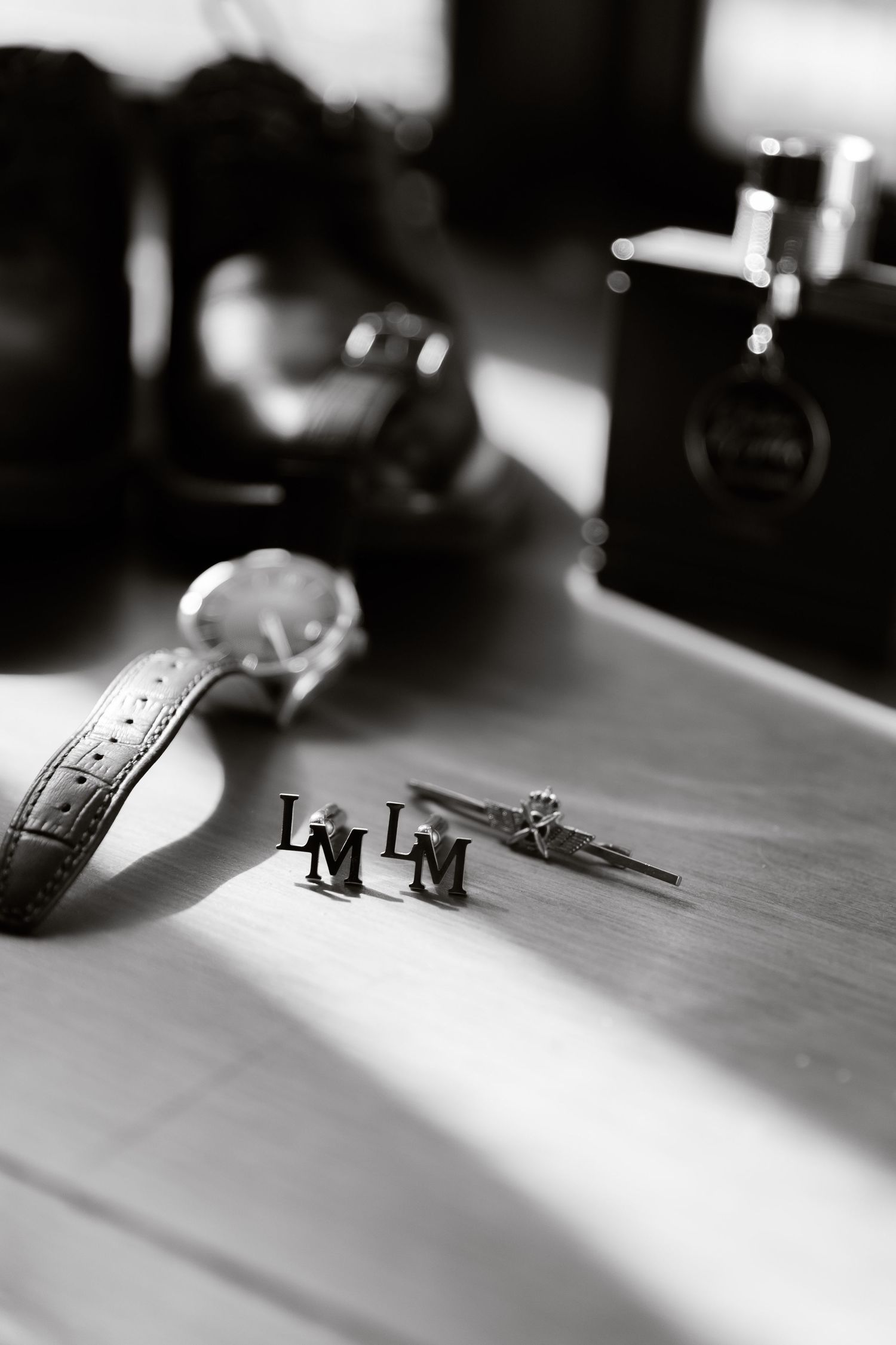 Black and white photo of personalized LM monogram cufflinks beside a watch and cologne on a flat surface.