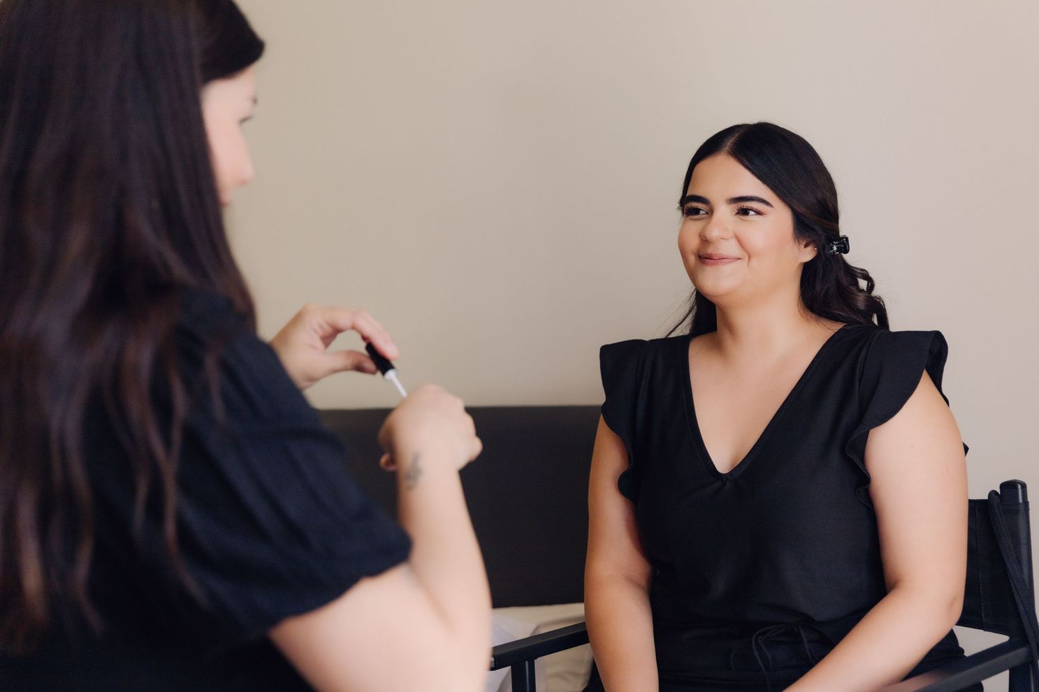 Makeup artist consulting with bride-to-be in black dress during wedding preparation in a bright room.