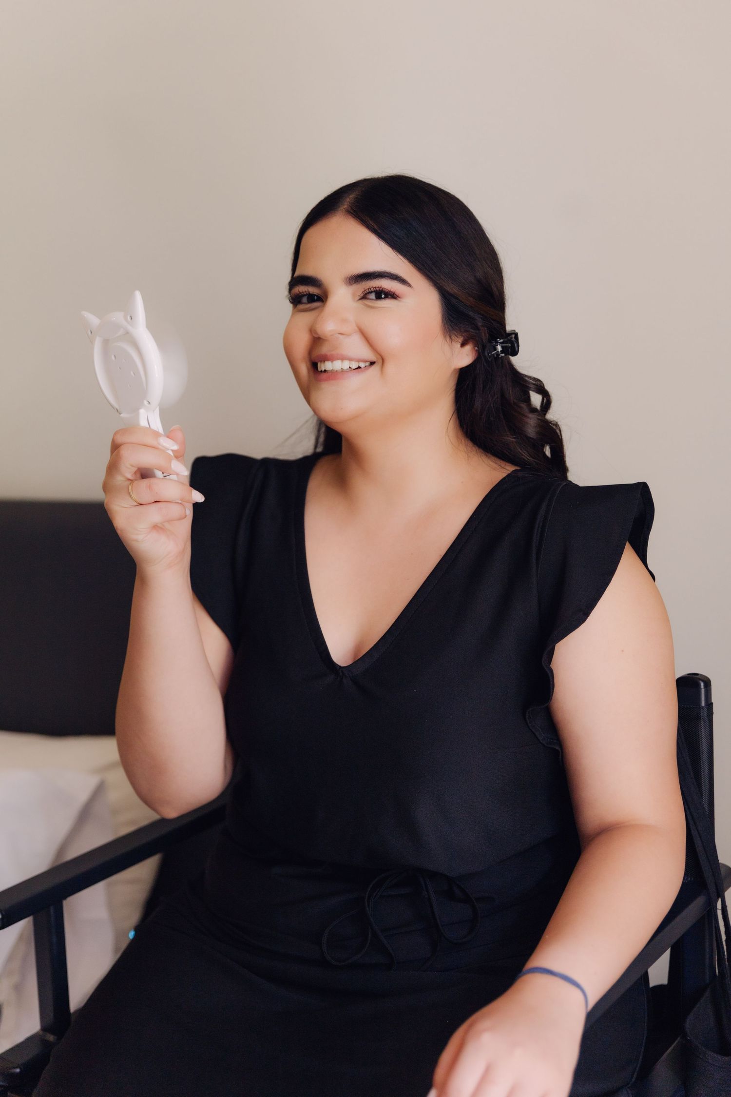 Smiling brunette woman in black dress holding white hair accessory while seated in styling chair.