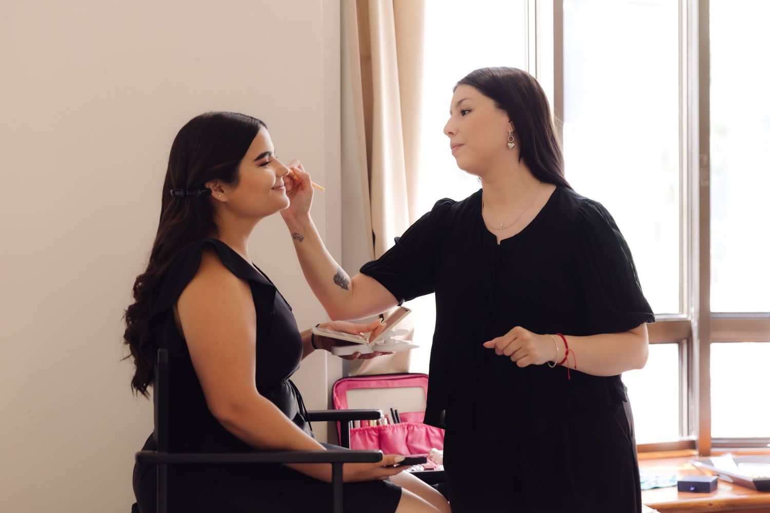 Professional makeup artist applying makeup to dark-haired woman in black dress at vanity station.