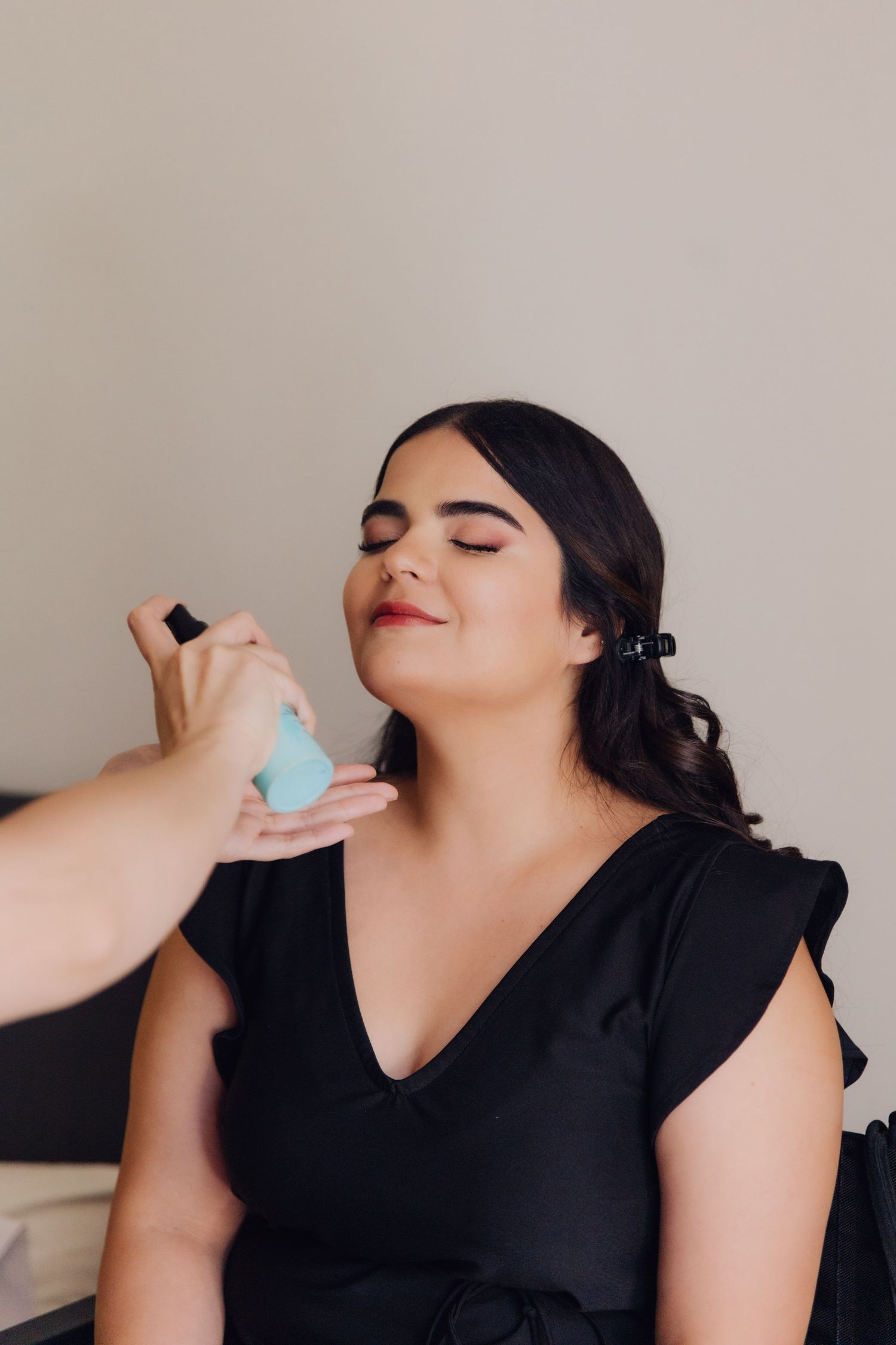 Makeup artist applying setting spray to woman in black dress with eyes closed during bridal prep.