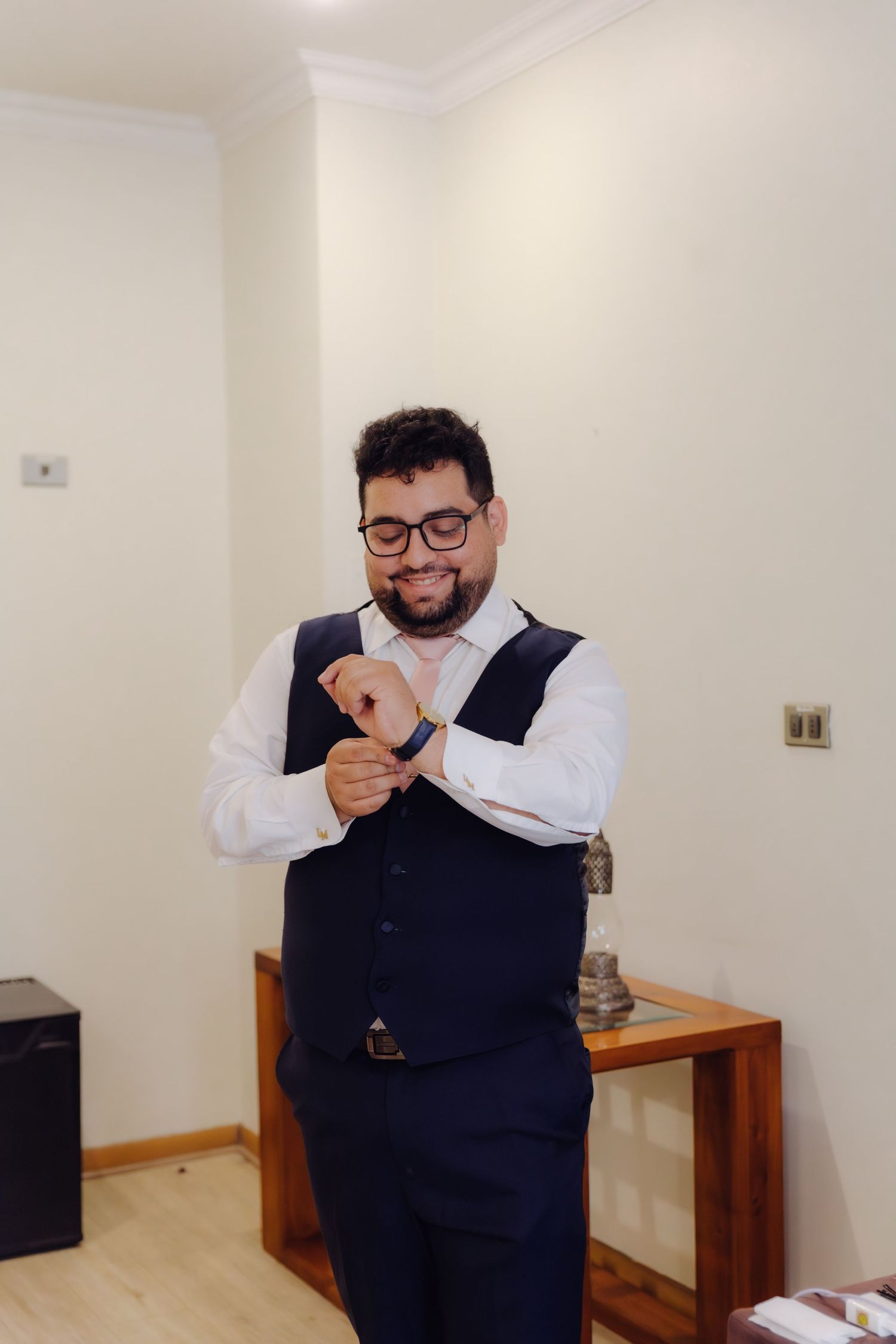 Smiling bearded groom in navy vest and white shirt fastening cufflinks while getting ready.