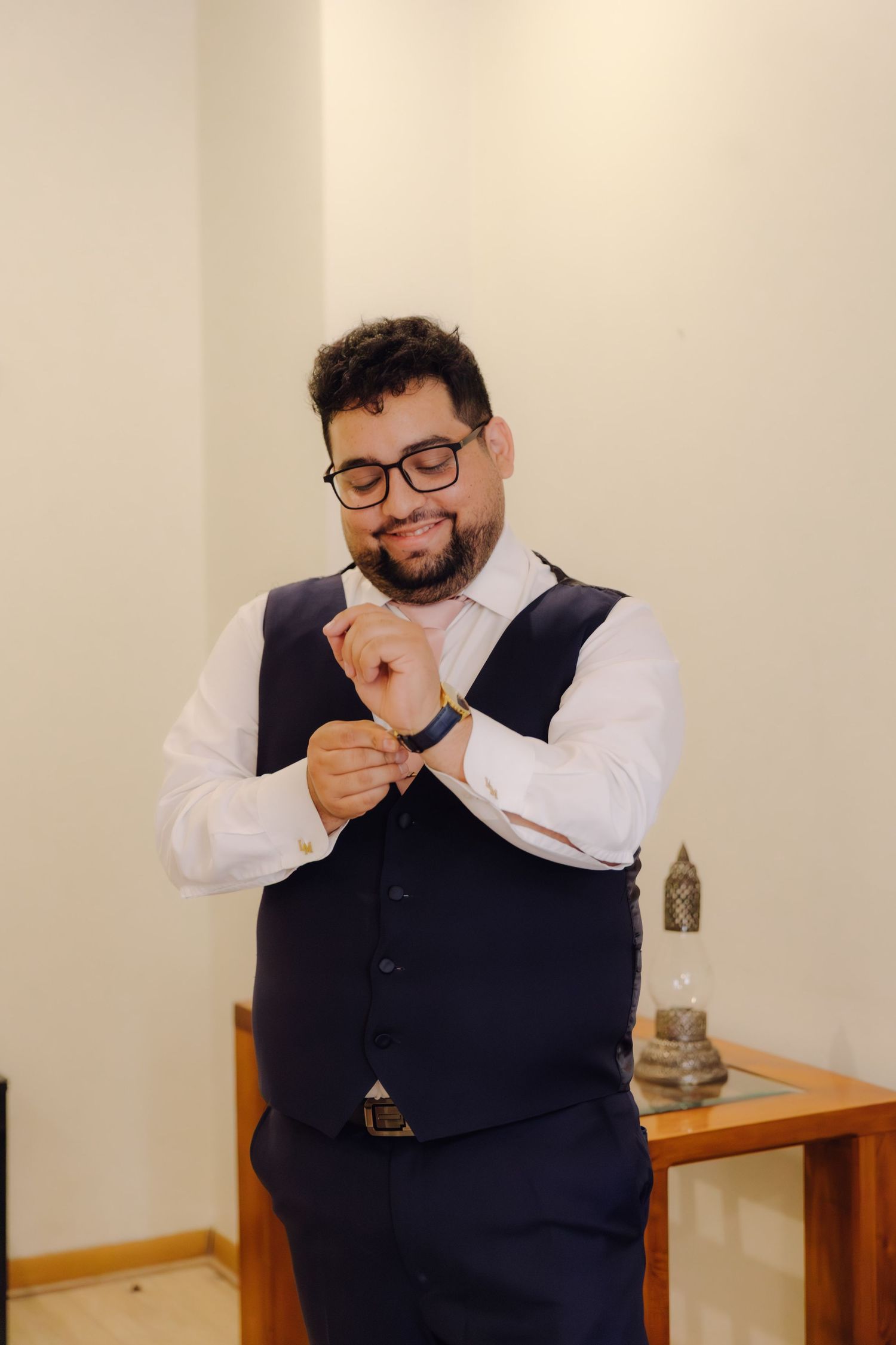 Handsome groom in navy waistcoat and glasses adjusting cufflinks while smiling during wedding prep.