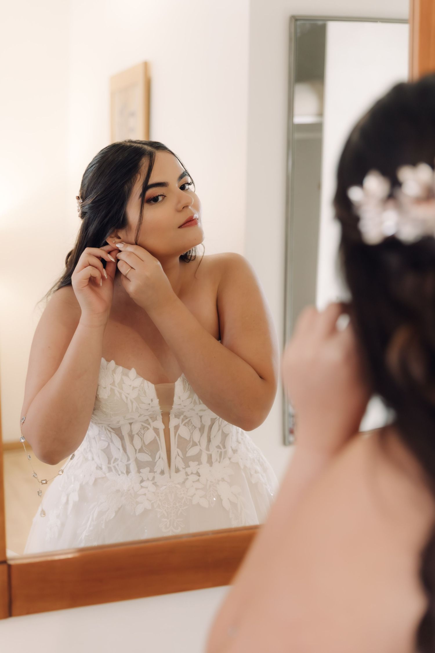 Beautiful bride in lace wedding gown putting on earrings while looking in mirror during preparation.