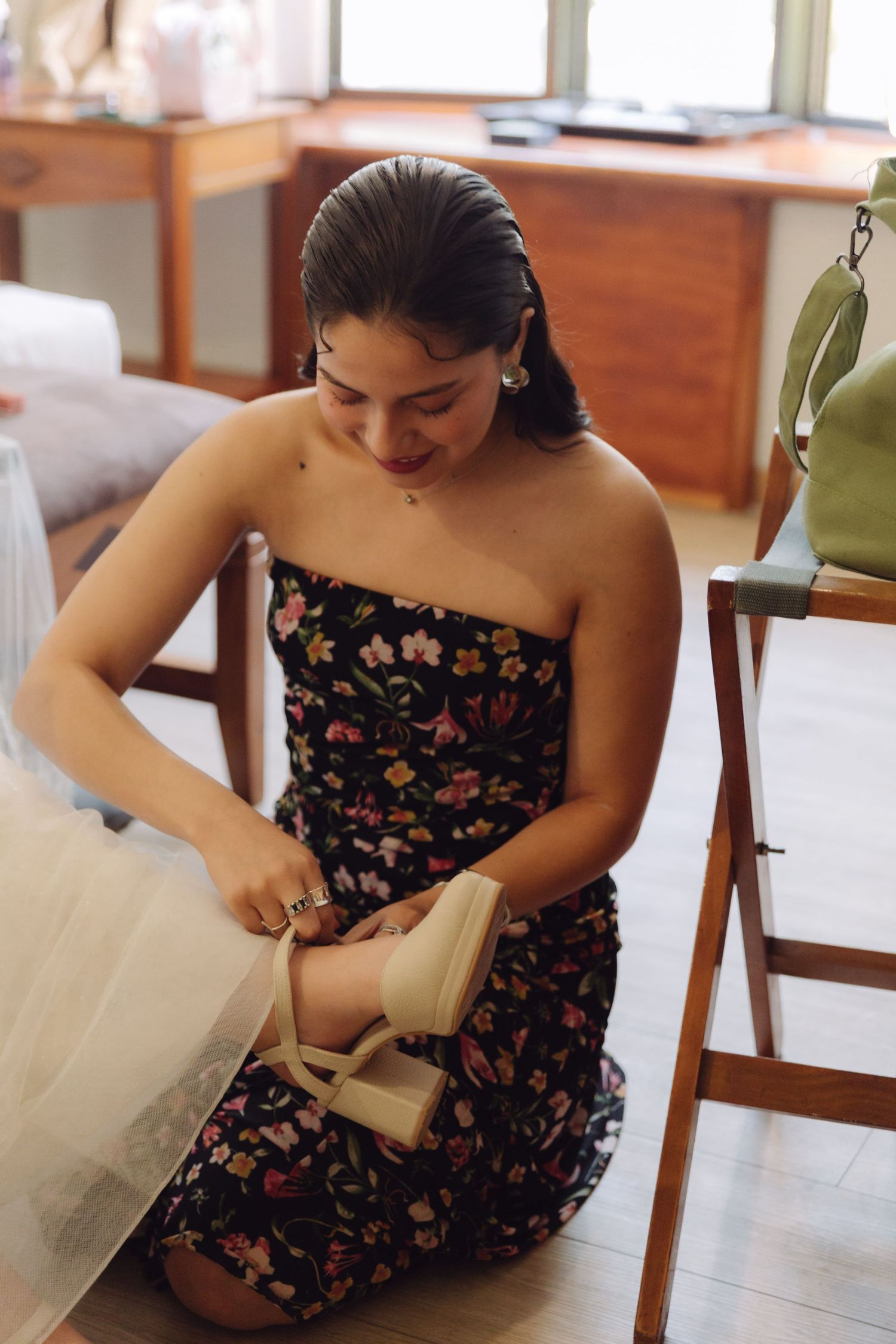 Woman in floral strapless dress kneeling to put on beige platform heels during wedding preparations.