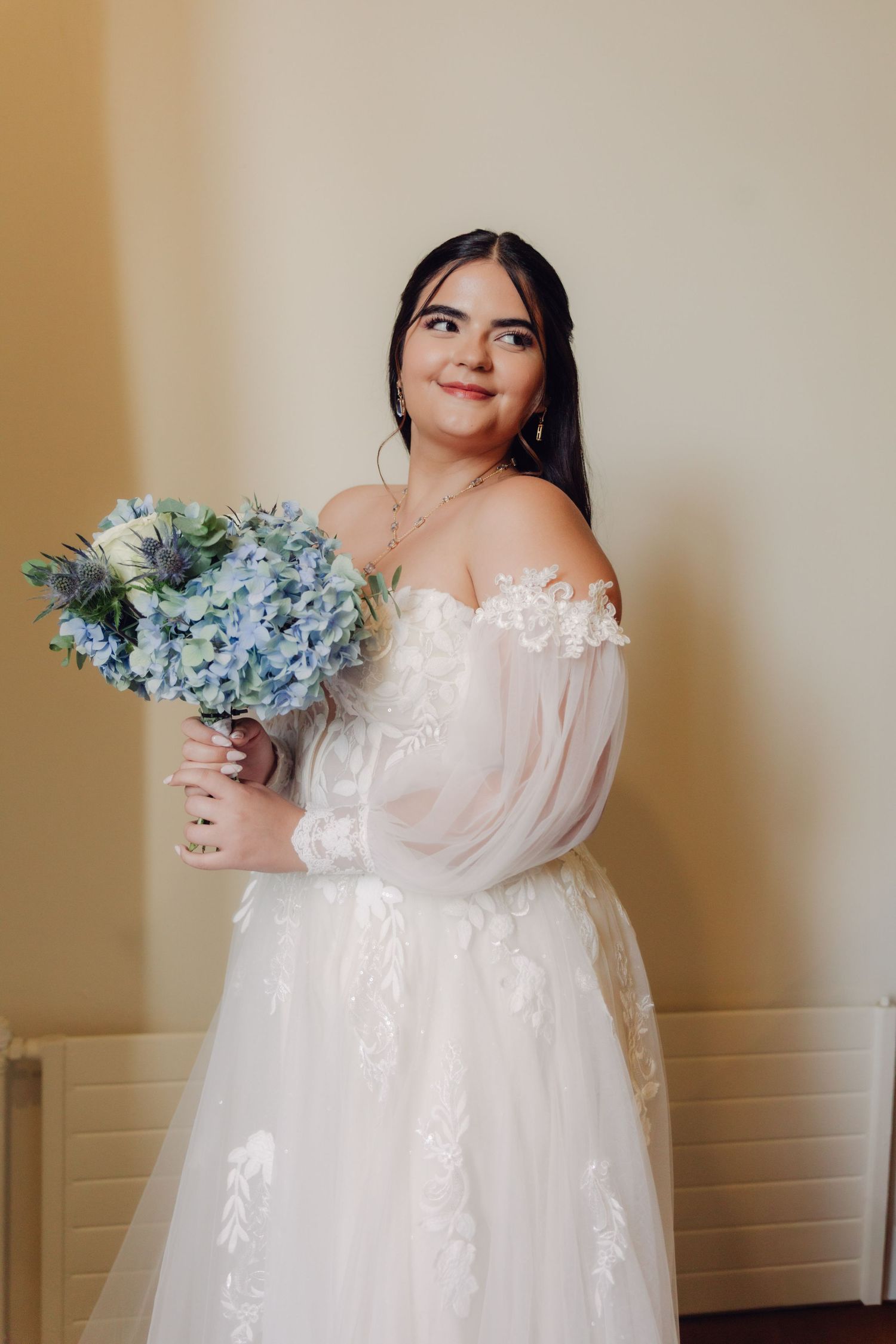 Radiant bride in off-shoulder lace wedding gown holding blue hydrangea bouquet indoors.