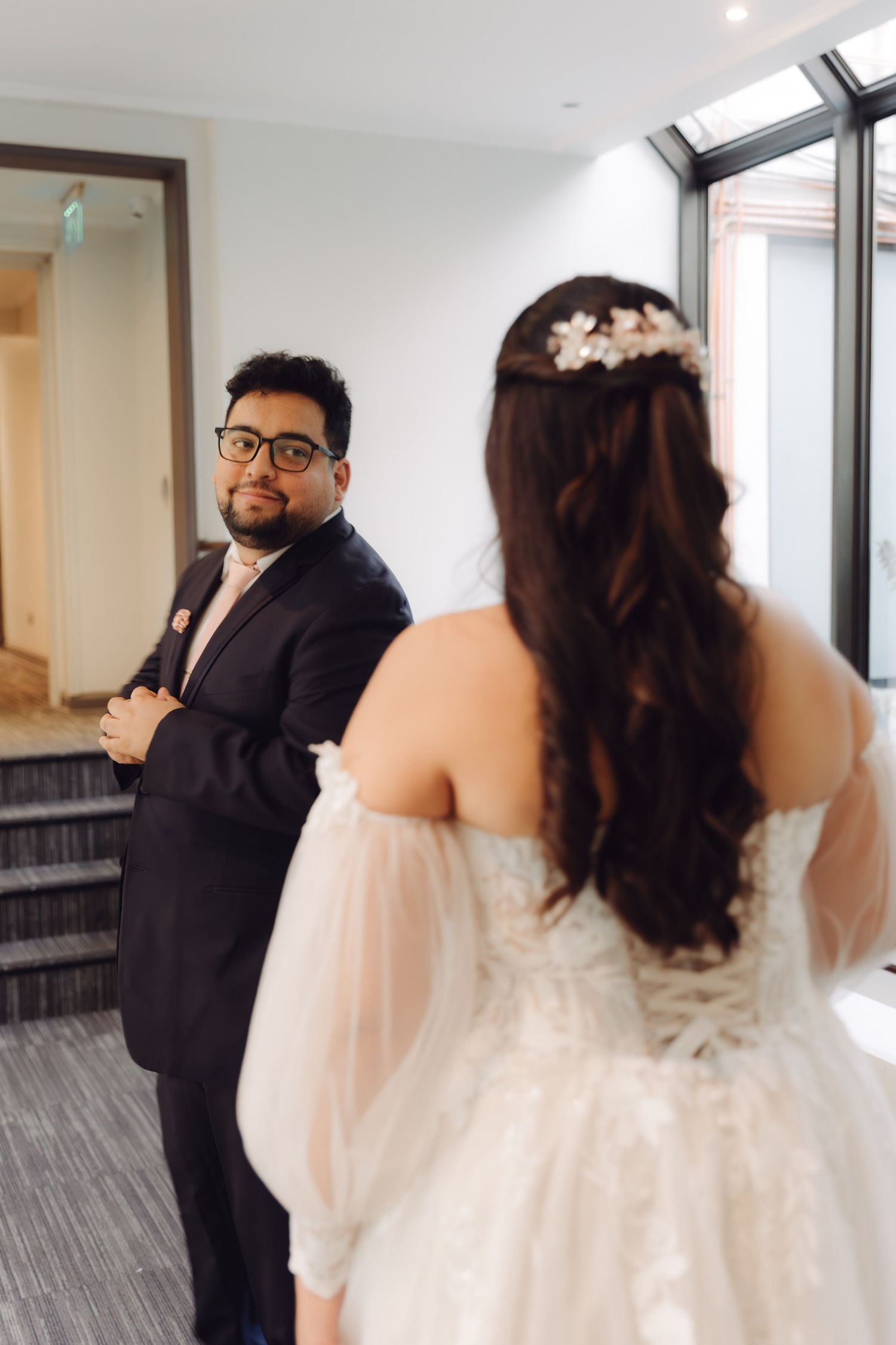 Groom in navy suit smiles at bride in off-shoulder wedding dress during first look moment indoors.