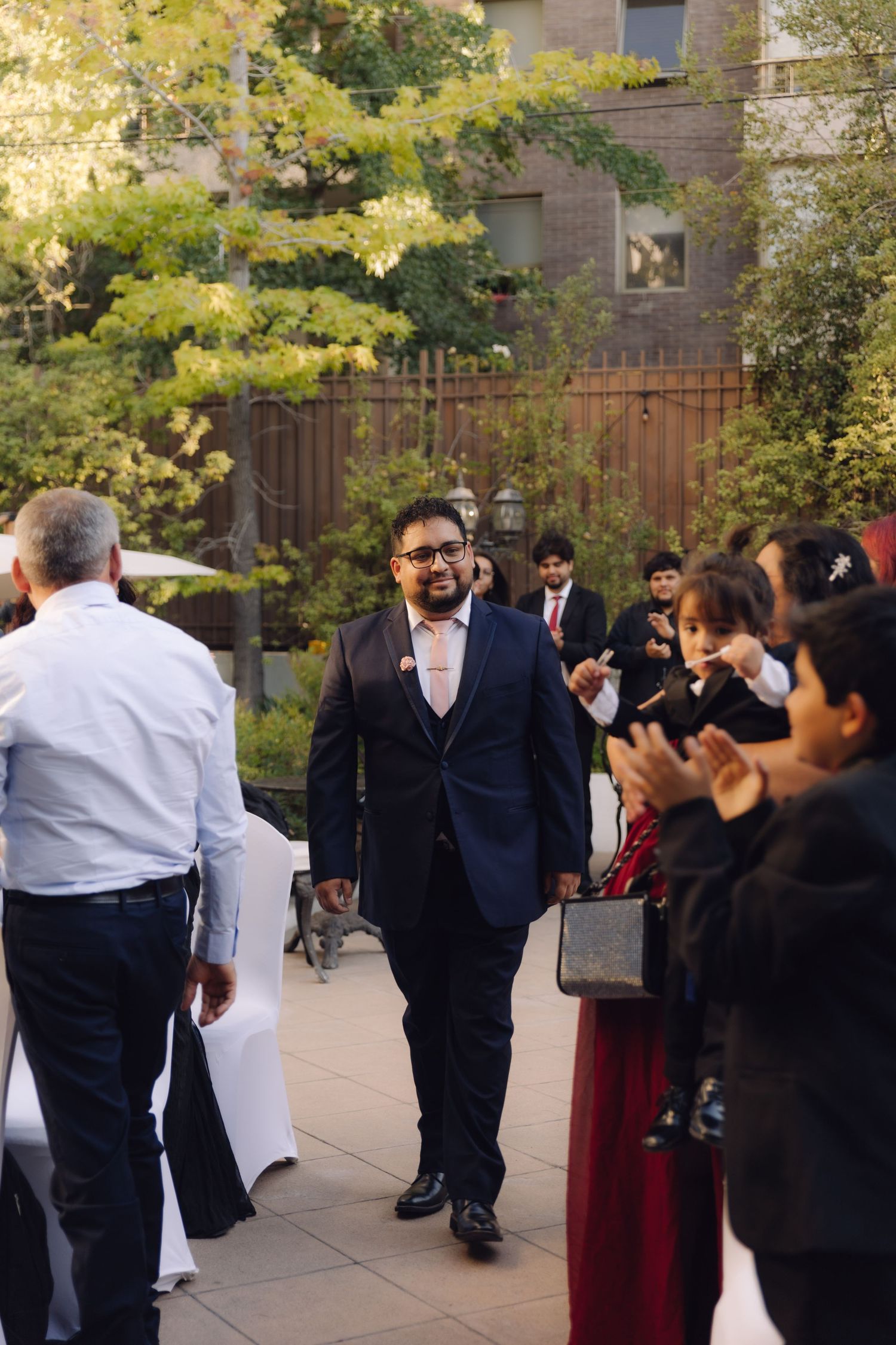 Groom in navy suit walks down outdoor aisle as wedding guests applaud and celebrate.
