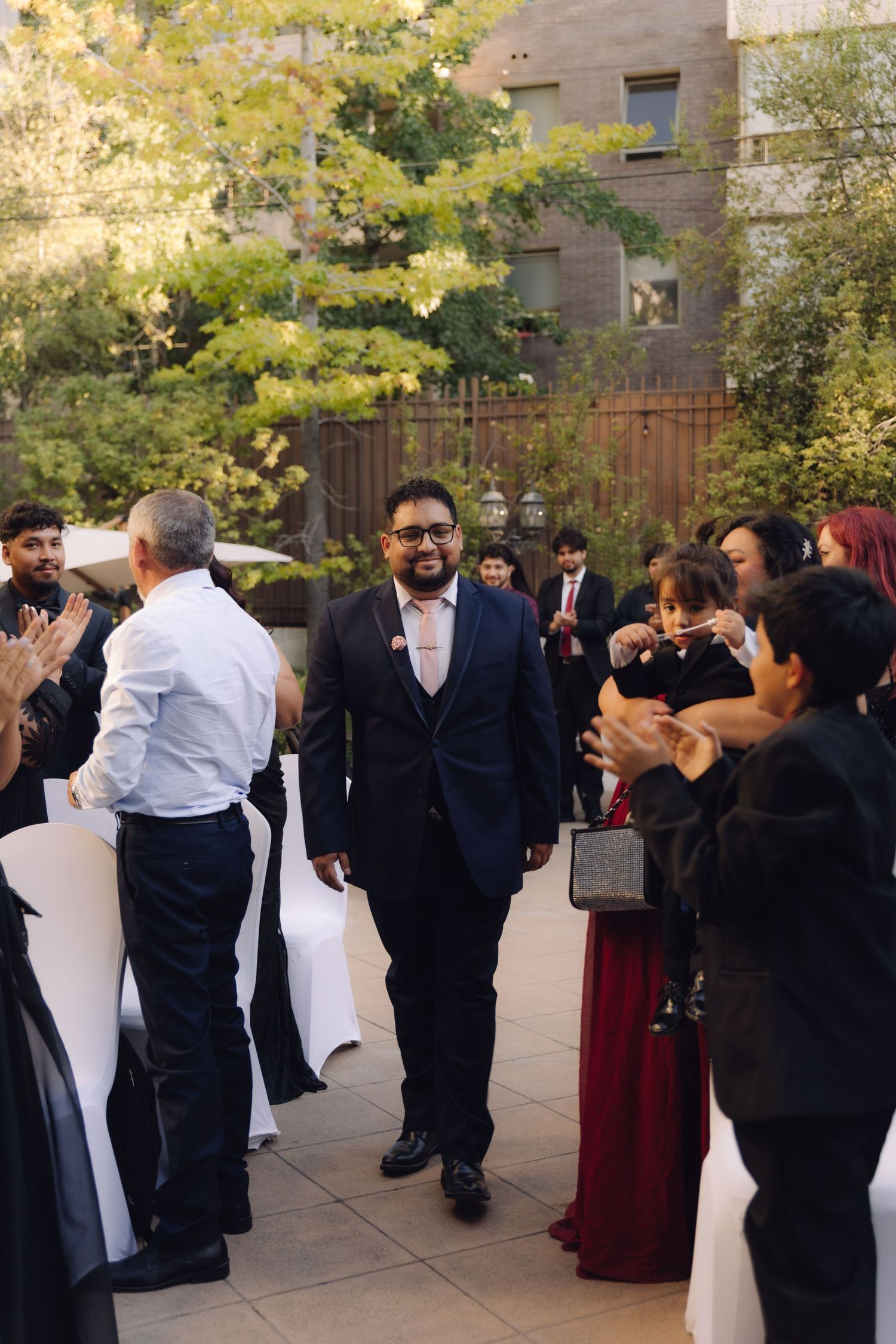Smiling groom in navy suit walks through applauding guests at outdoor wedding ceremony.
