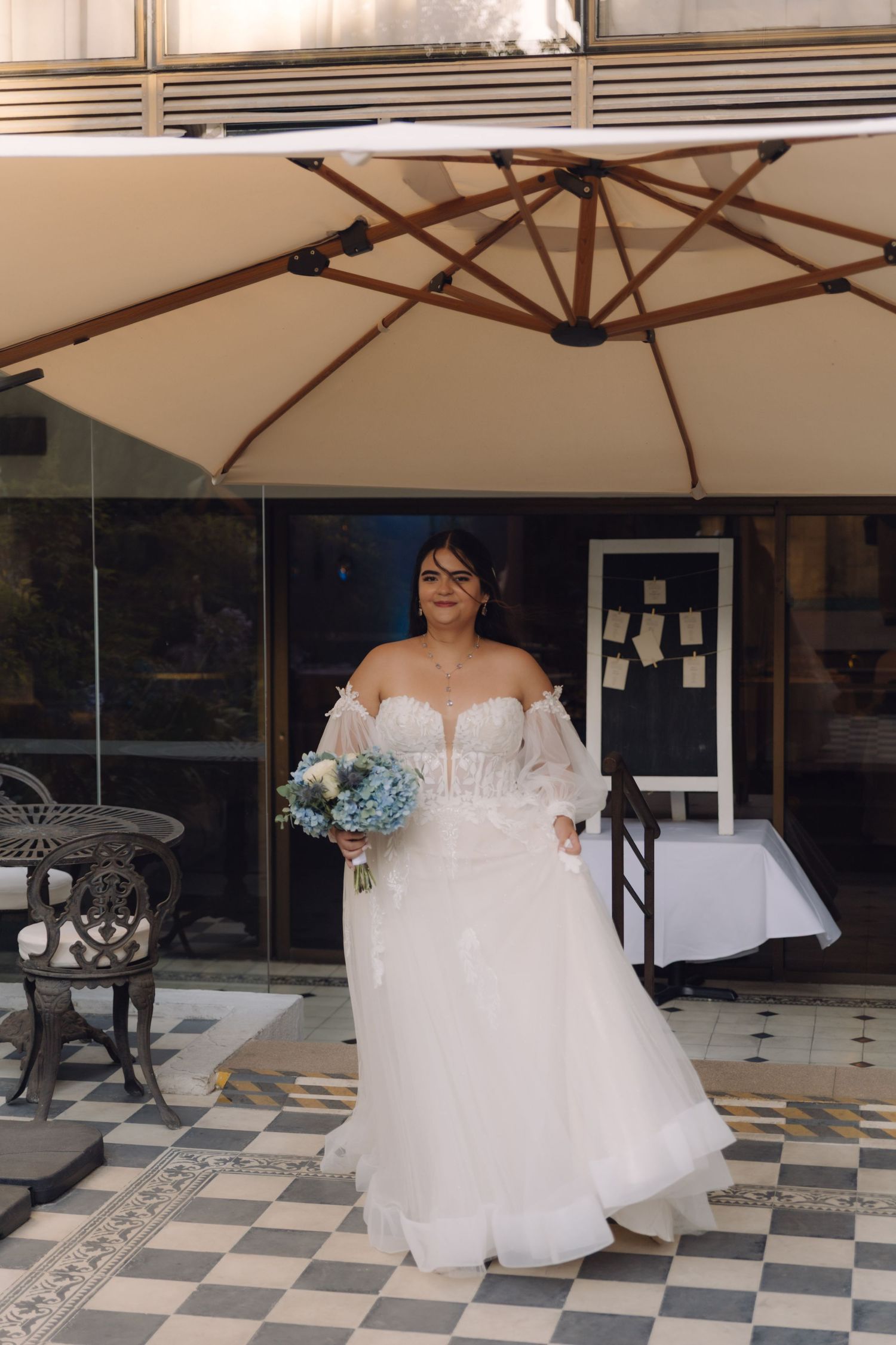 Bride in off-shoulder white wedding gown holds blue hydrangea bouquet under outdoor patio umbrella.