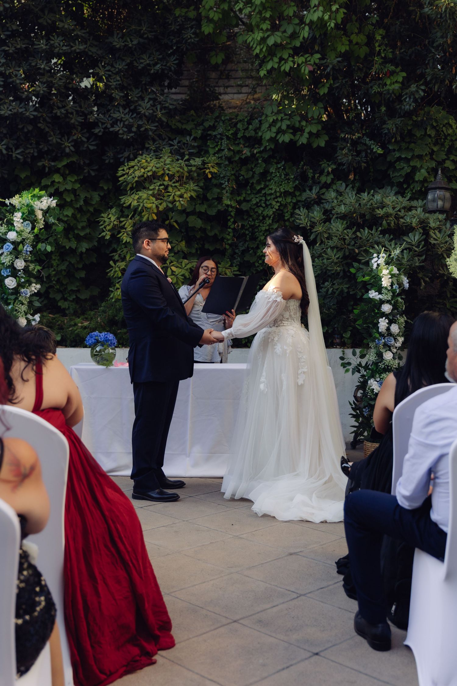 Bride and groom exchange vows at outdoor ceremony before lush green floral arch backdrop.