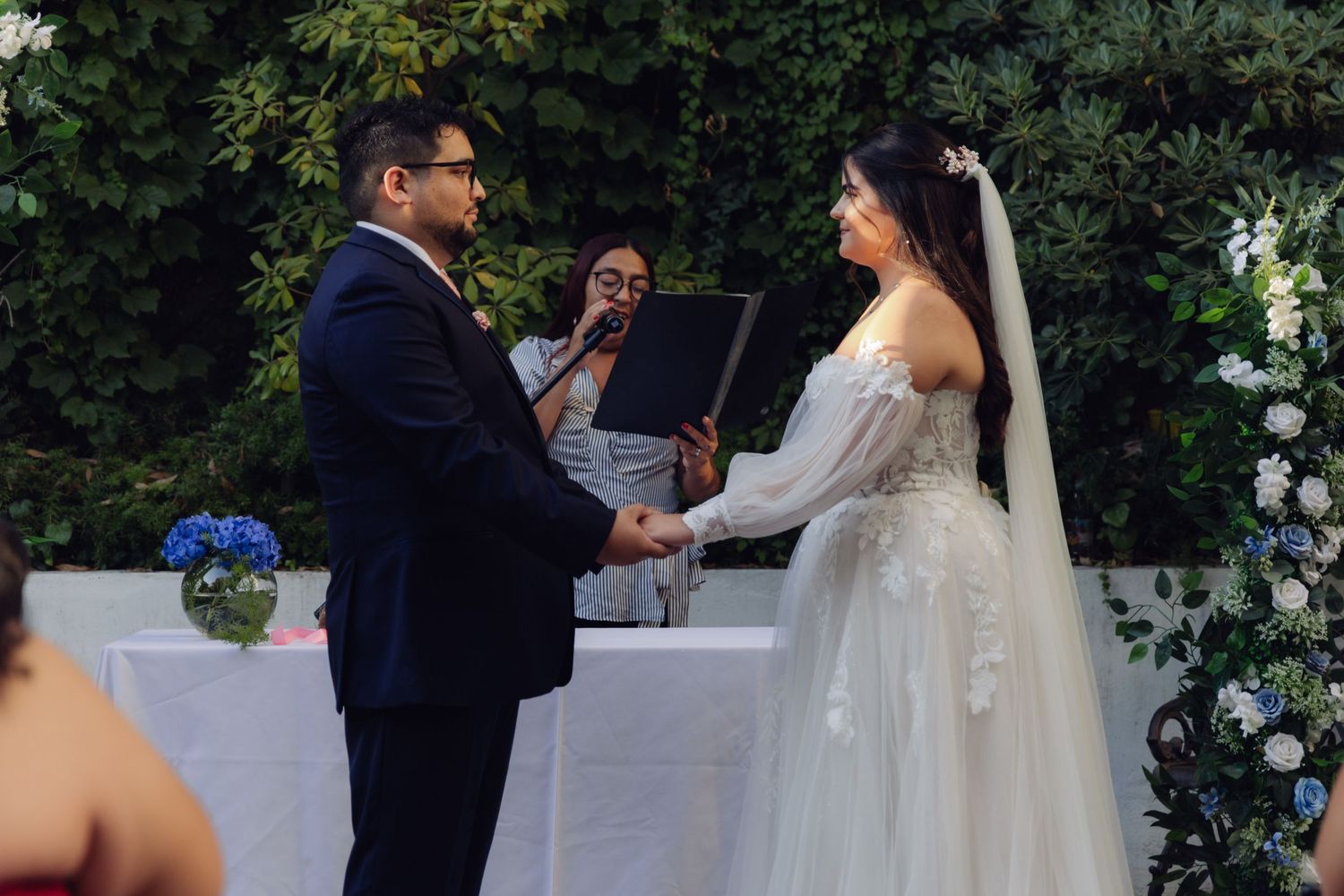 Couple holds hands exchanging wedding vows as officiant reads ceremony script outdoors.