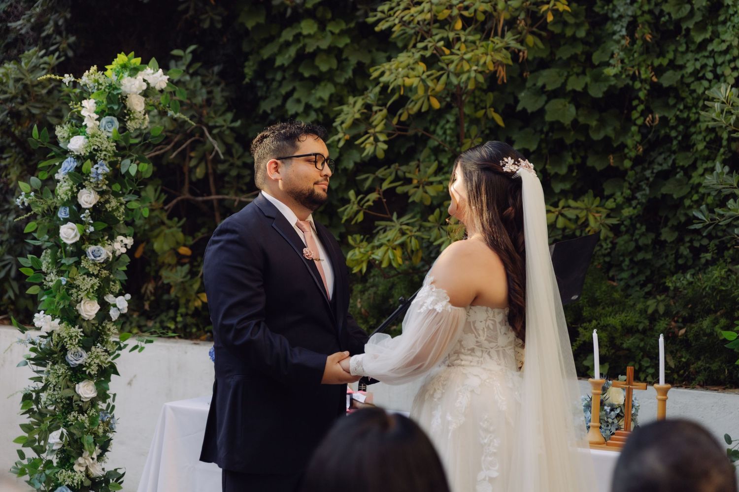 Bride in white veil and groom in navy suit hold hands at floral arch altar.