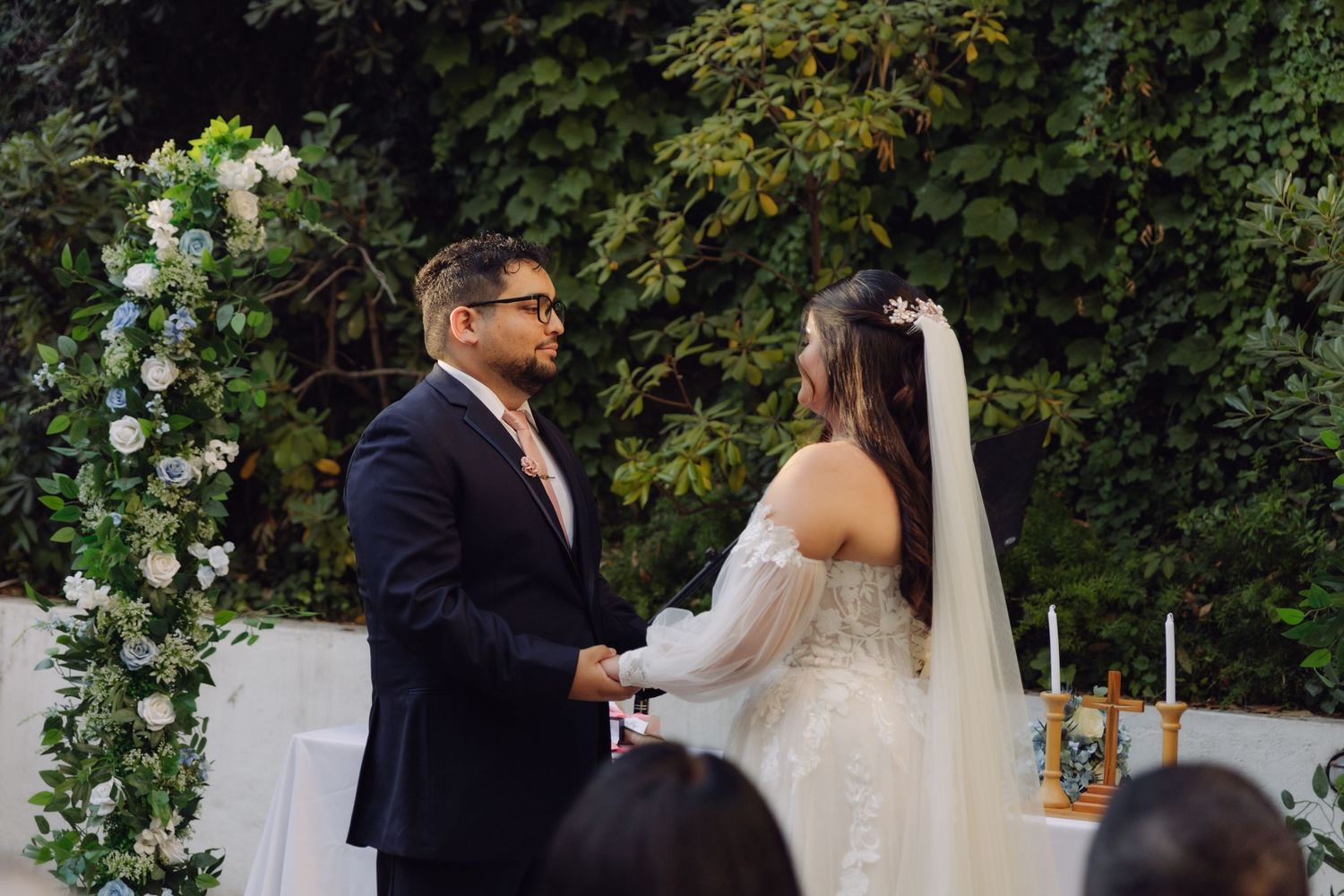 Bride and groom face each other holding hands during outdoor wedding ceremony at floral arch.