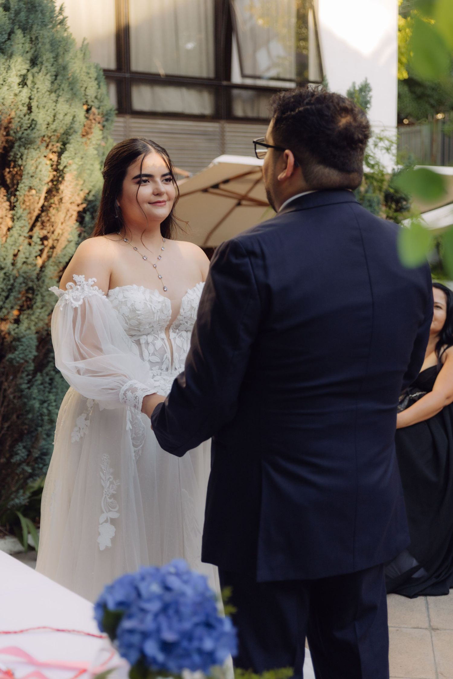 Bride in lace wedding gown holds hands with groom in navy suit during outdoor wedding ceremony.