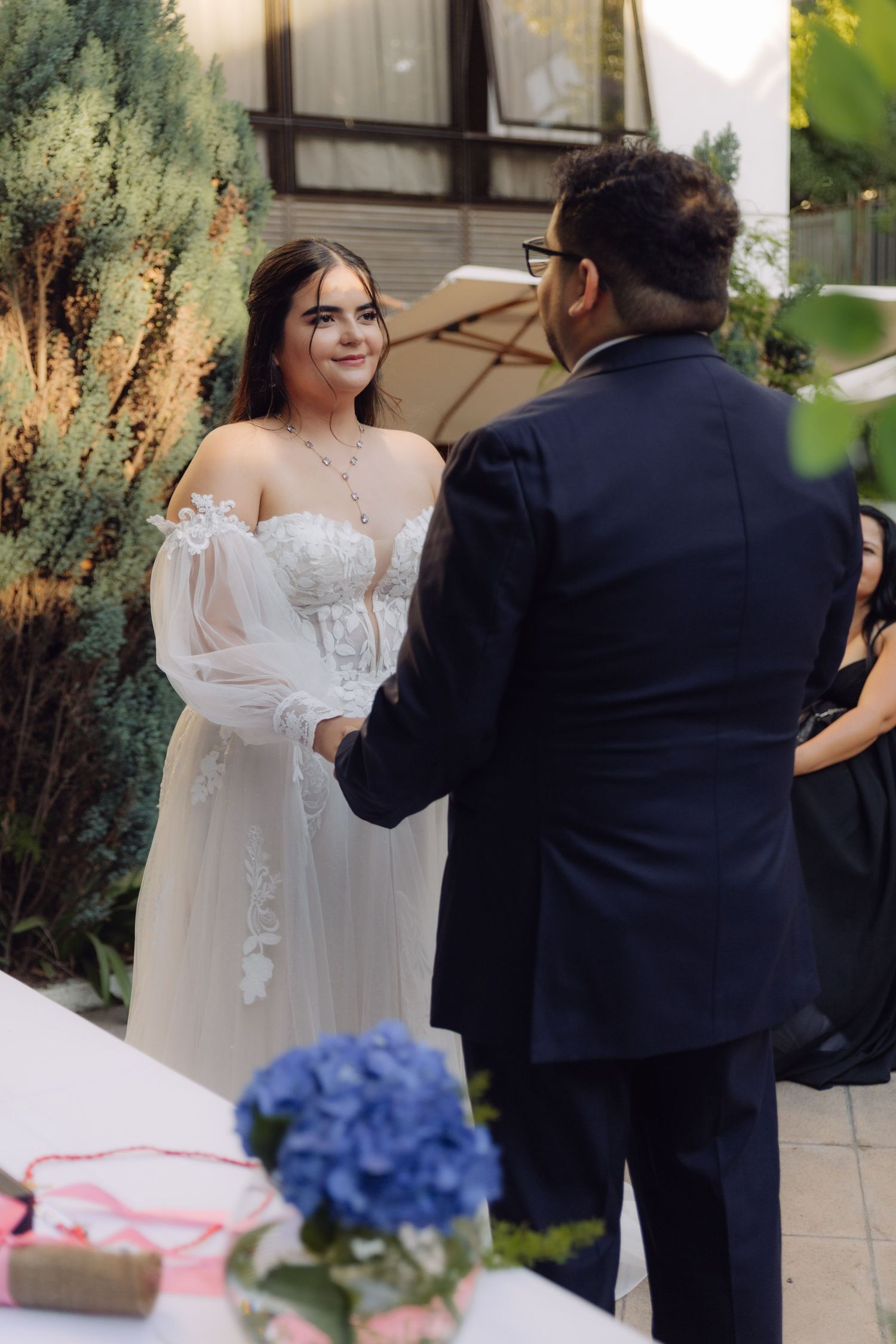 Smiling bride in off-shoulder lace gown exchanges vows with groom at outdoor garden wedding.