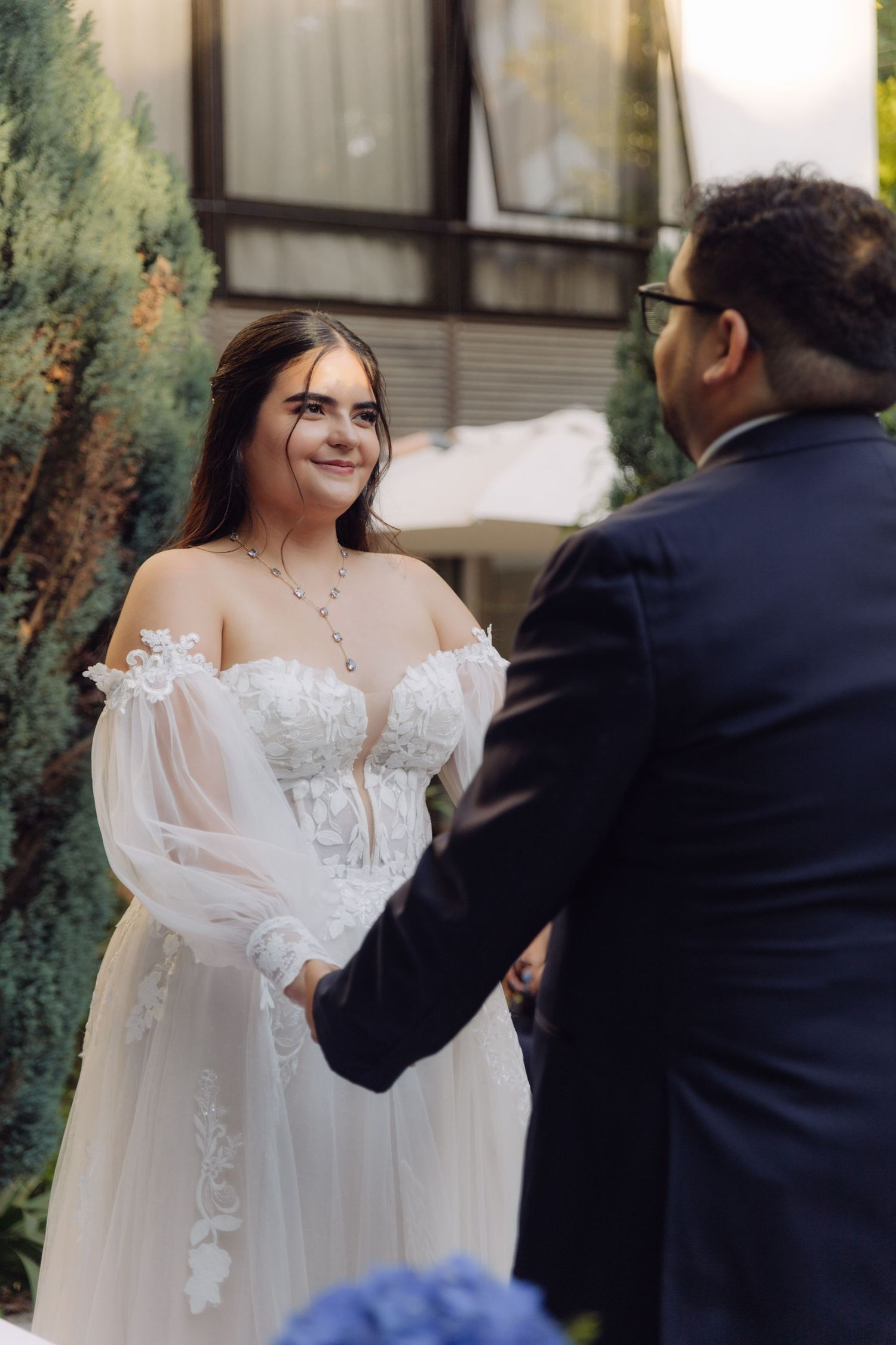 Bride in floral lace wedding dress holds hands with groom in dark suit at outdoor ceremony.