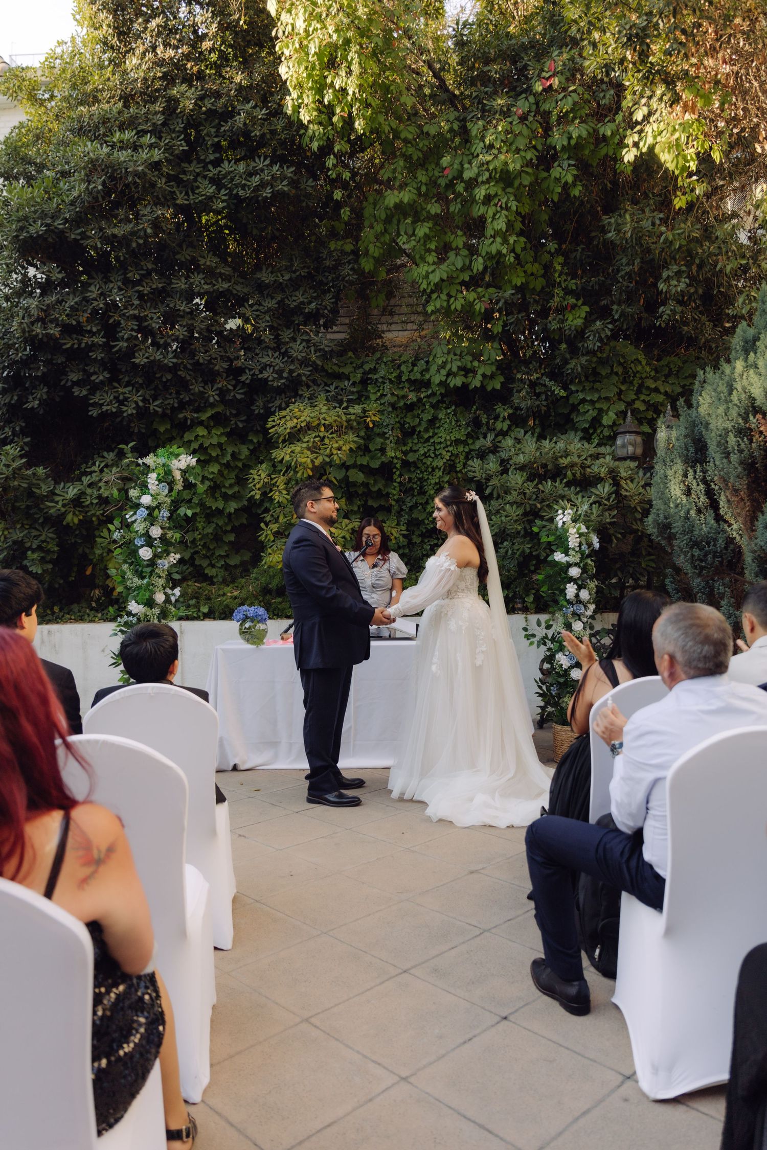 Couple exchanges vows at outdoor garden wedding ceremony with guests seated in white chairs.