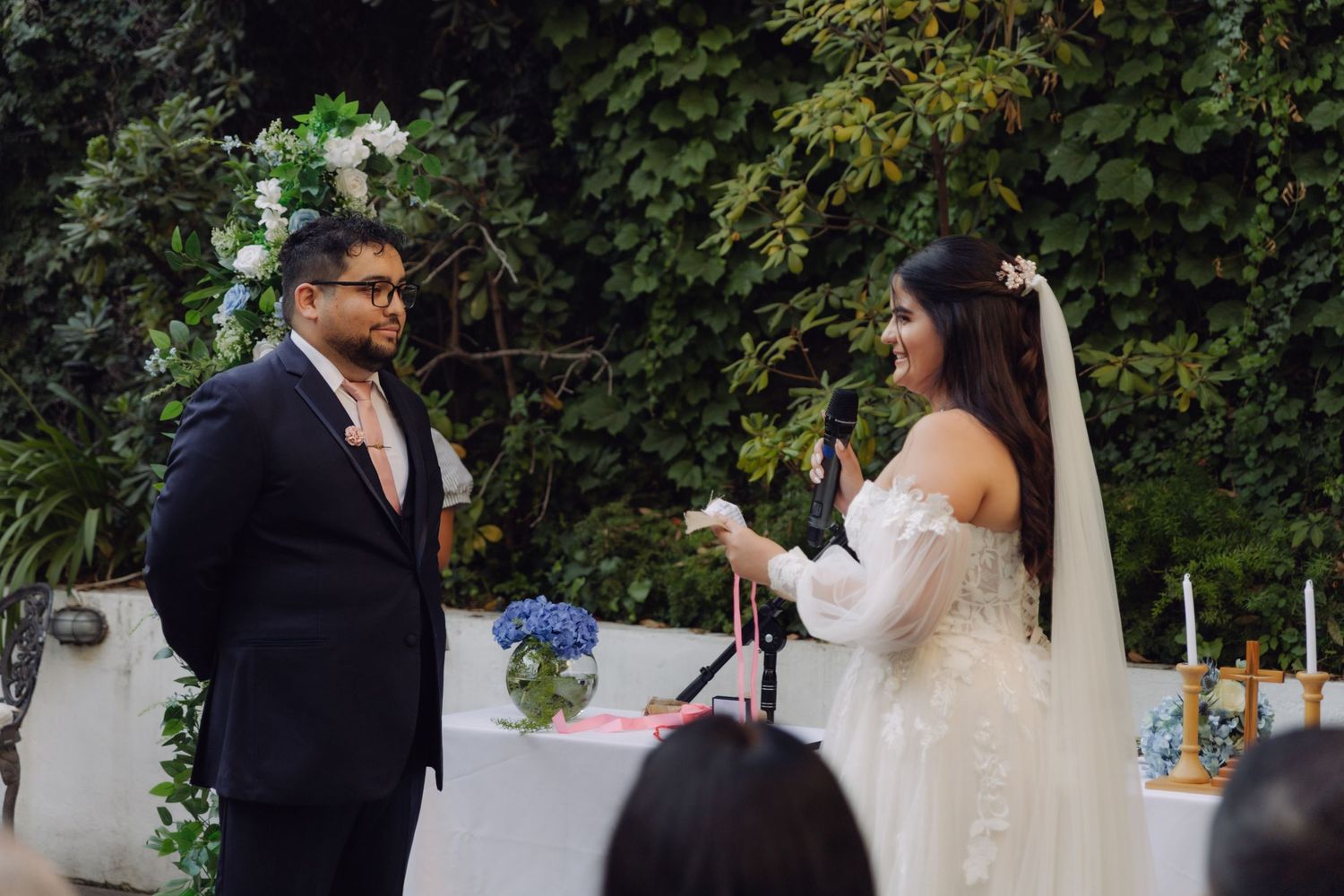 Bride reads vows into microphone while groom listens at outdoor garden wedding ceremony.