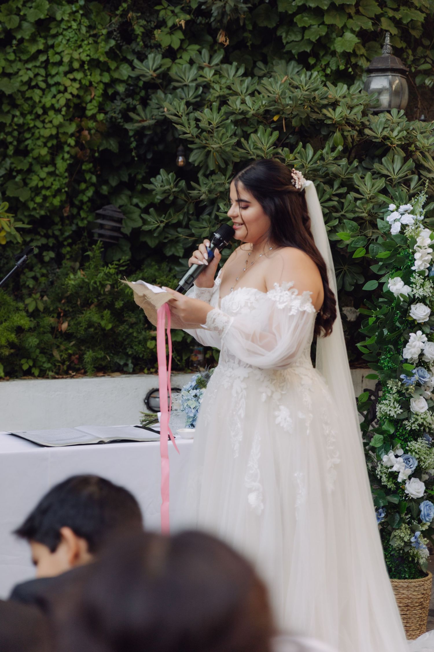 Bride in lace gown and veil reads wedding vows into microphone at outdoor garden ceremony.